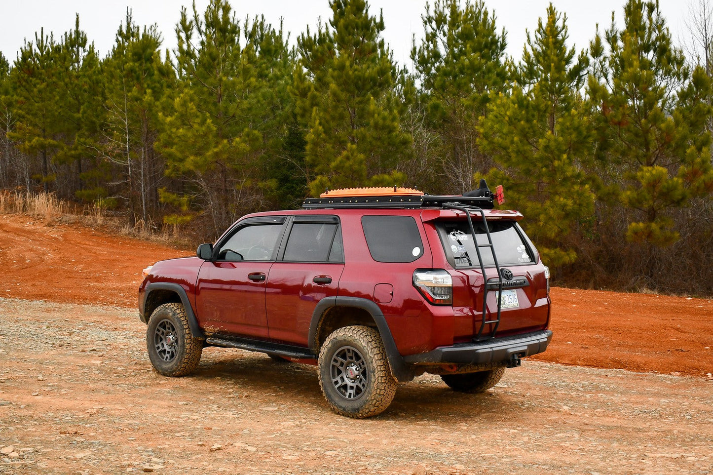 A 5th Gen 4Runner with a 3/4 Roof Rack is parked on a dirt road, equipped with off-road tires and ruggedized crossbars, surrounded by green pine trees and reddish soil beneath an overcast sky.