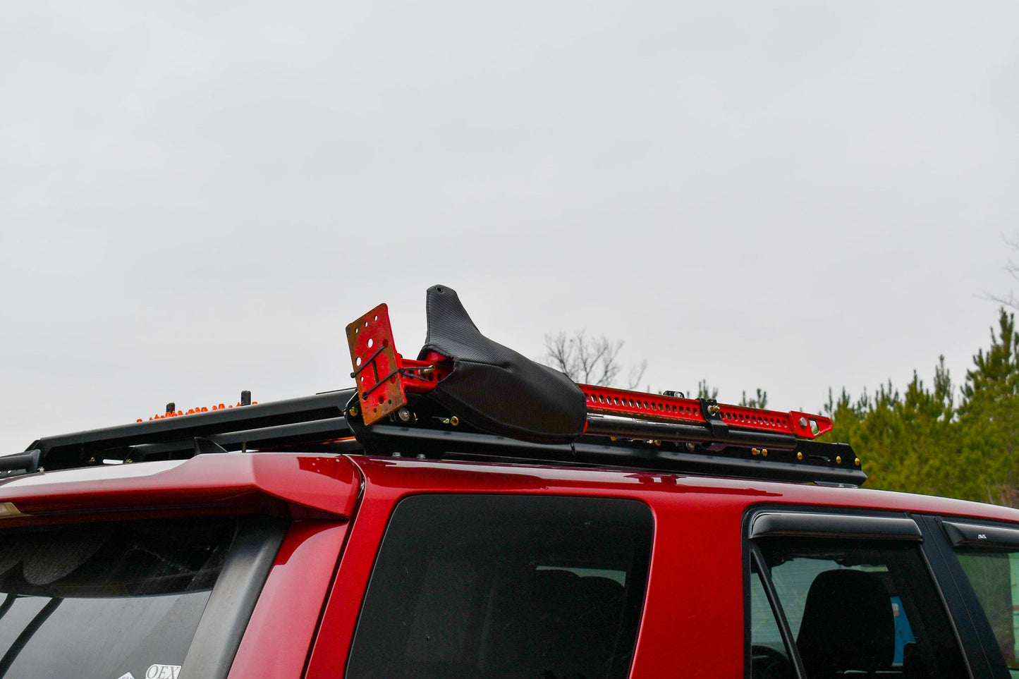 A 5th Gen 4Runner with a 3/4 Roof Rack carries a black seat and red metal jack, parked outdoors under cloudy skies and trees—showcasing the practicality of the 3/4 Roof Rack - 5th Gen 4Runner in real-world use.
