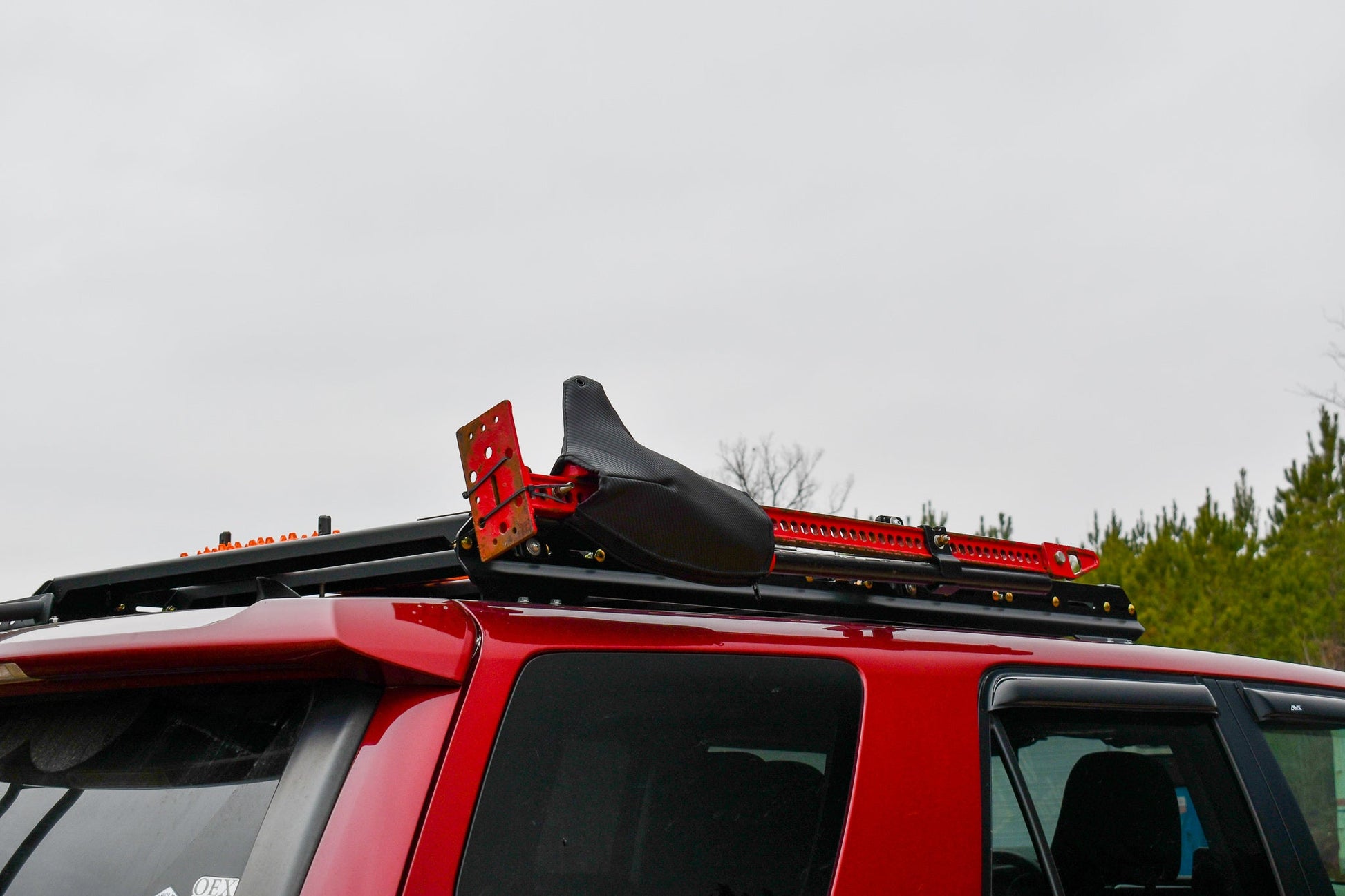 A 5th Gen 4Runner with a 3/4 Roof Rack carries a black seat and red metal jack, parked outdoors under cloudy skies and trees—showcasing the practicality of the 3/4 Roof Rack - 5th Gen 4Runner in real-world use.