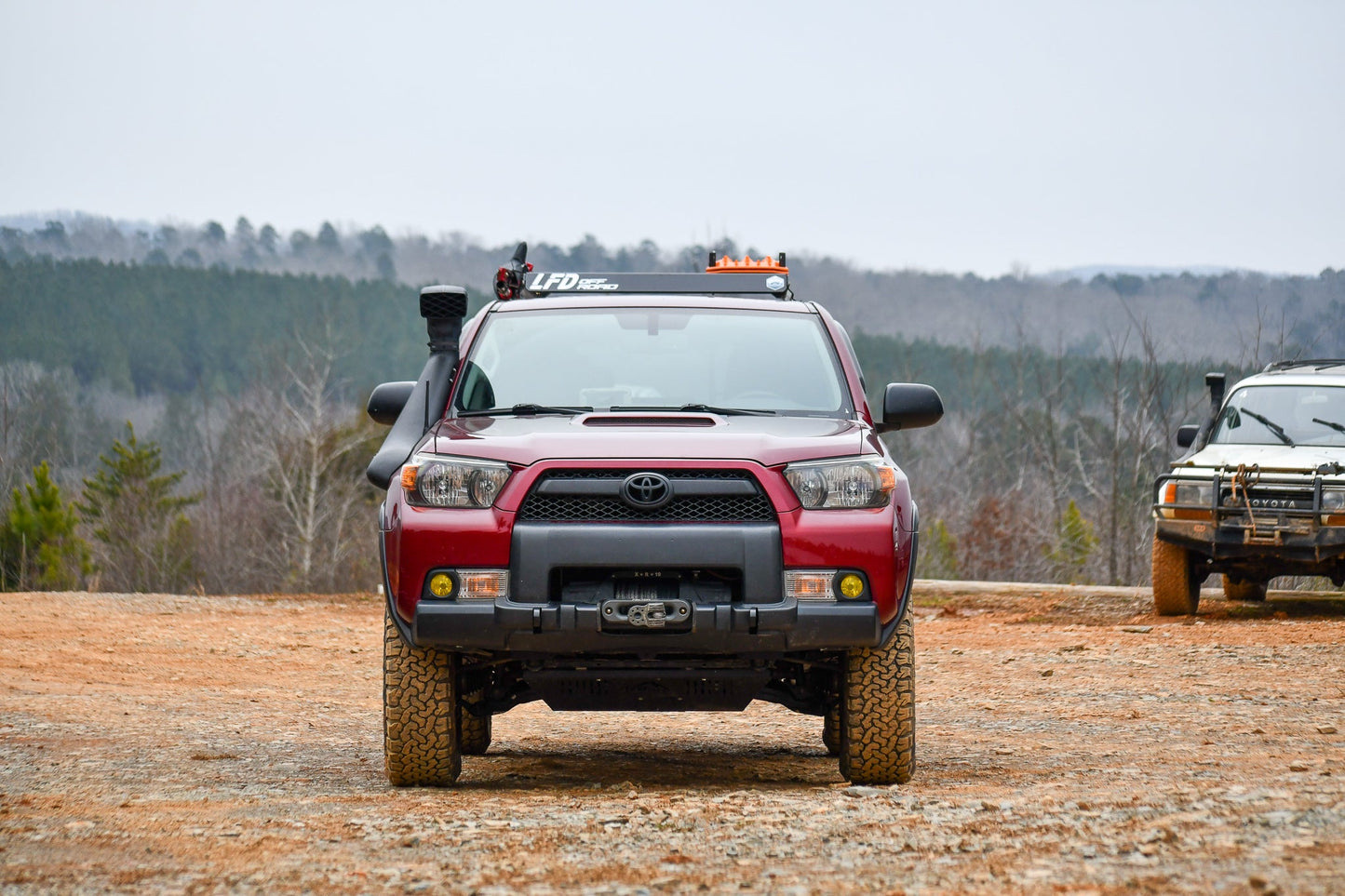 A red Toyota 5th Gen 4Runner with a 3/4 Roof Rack is parked on rocky terrain, facing forward. Another off-road vehicle is partially visible on the right, set against a forested landscape under cloudy skies.