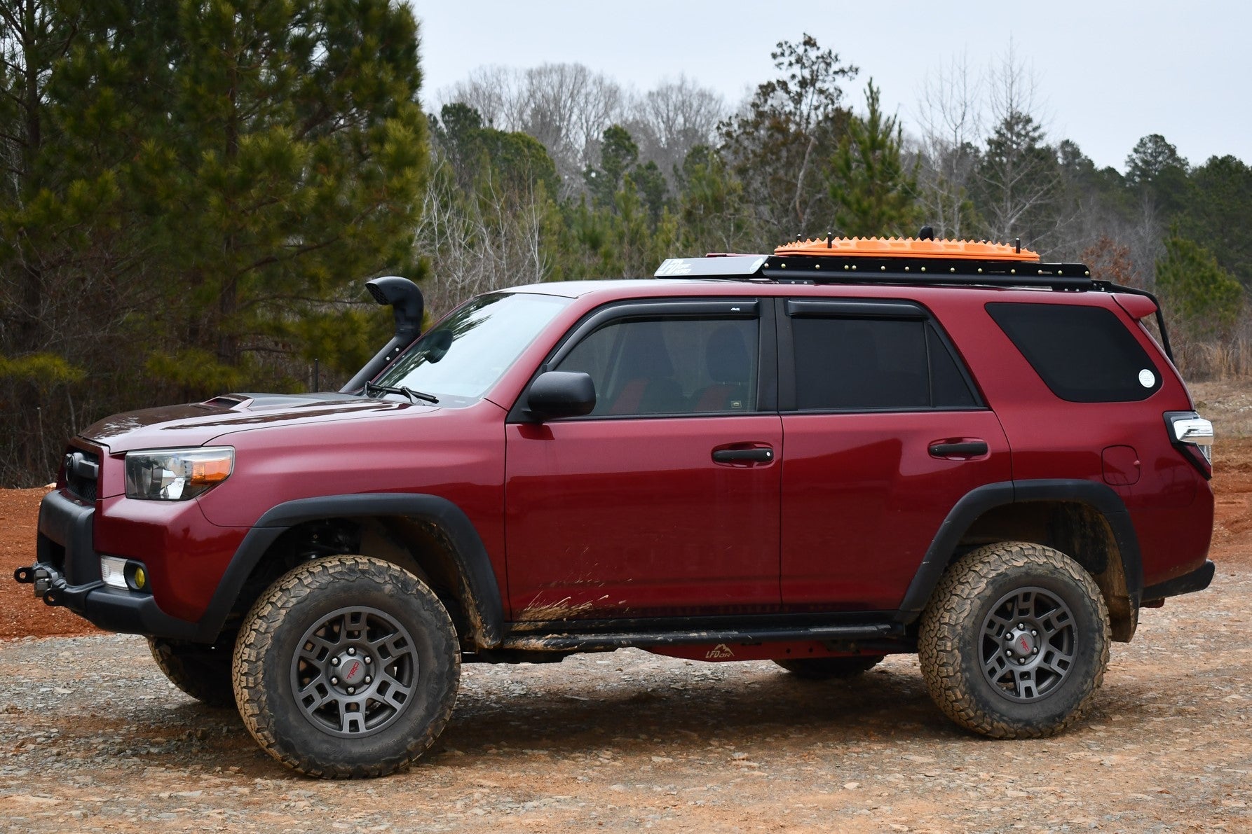 A red 5th Gen 4Runner with rugged crossbars, off-road tires, muddy lower panels, and a 3/4 Roof Rack - 5th Gen 4Runner is parked on a dirt path amid trees under overcast skies.