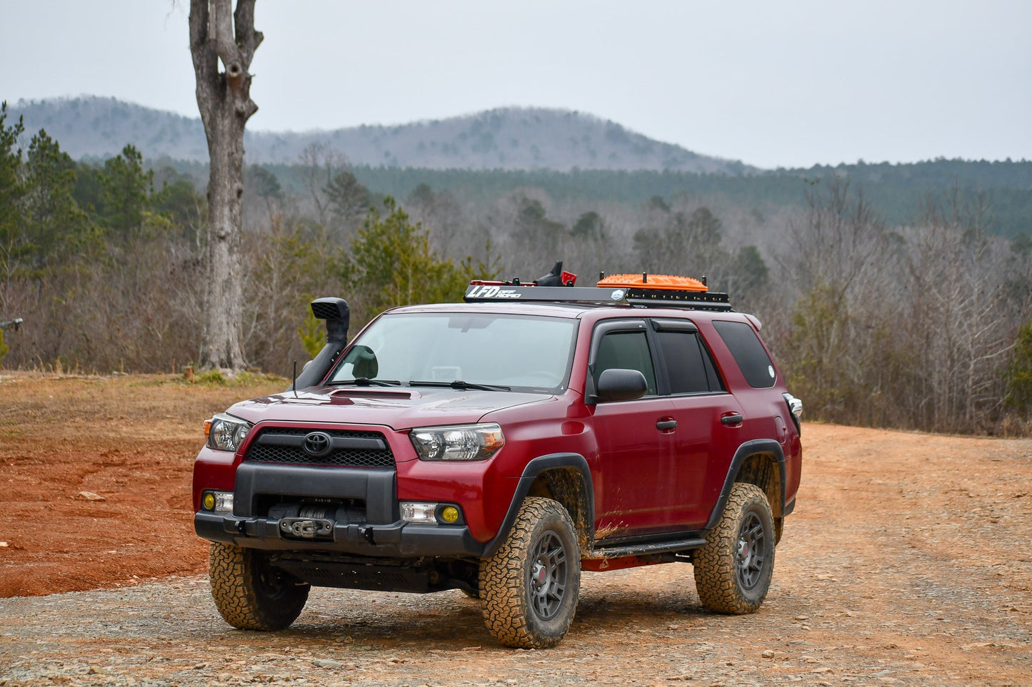 A red Toyota 4Runner with a 3/4 Roof Rack for the 5th Gen model is parked on a dirt road in a forested, mountainous area under an overcast sky.