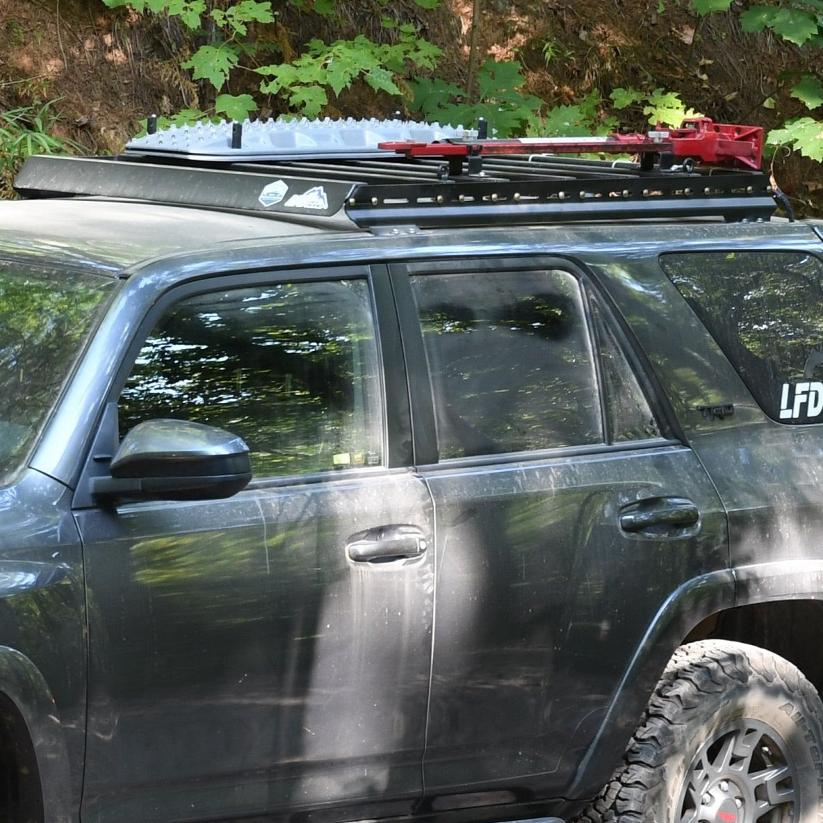 A dark SUV, the 5th Gen 4Runner, is parked outdoors with off-road tires and a 3/4 Roof Rack holding recovery boards and a red hi-lift jack, set against green foliage.