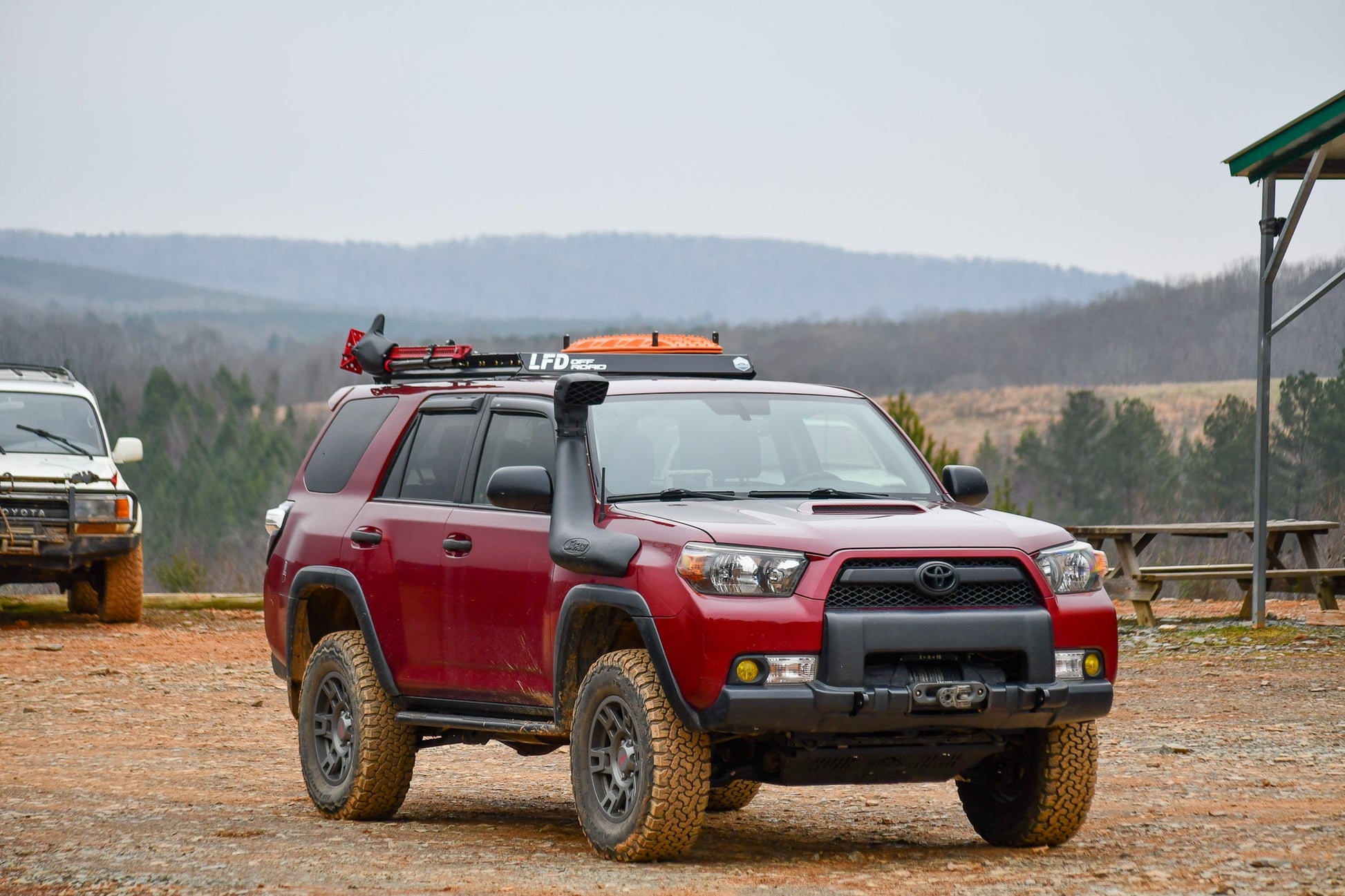 A red lifted Toyota 4Runner with a 3/4 Roof Rack - 5th Gen 4Runner and off-road tires is parked on dirt before distant hills, with another SUV partly visible in the background and rack accessories mounted on top.