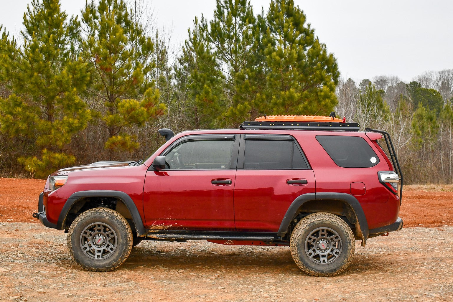 A 5th Gen 4Runner with a 3/4 Roof Rack is parked on a dirt and gravel road, surrounded by pine trees under a cloudy sky.