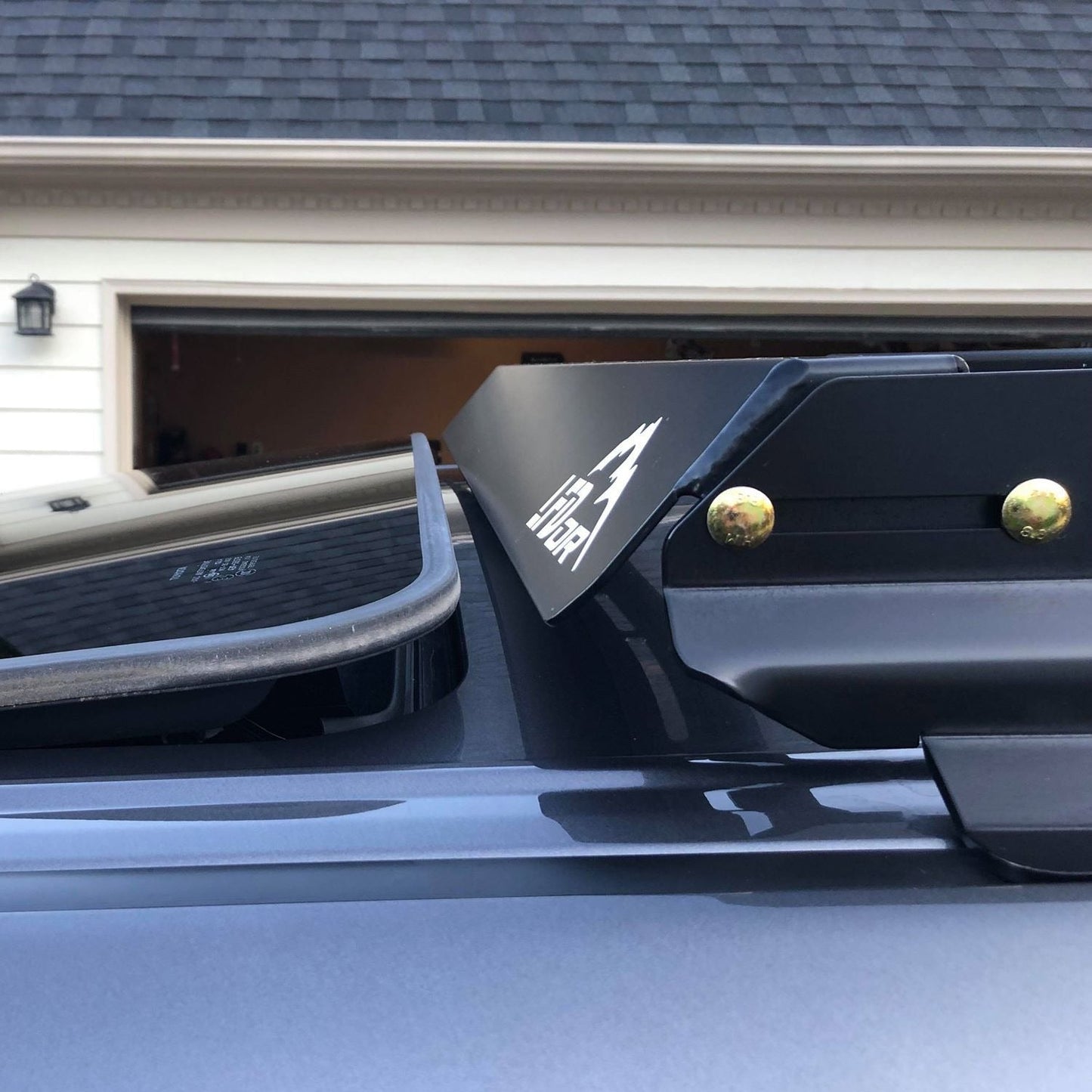 Close-up of a 3/4 Roof Rack - 5th Gen 4Runner in black with the Viper logo and rugged crossbars, mounted on a gray car parked in front of a house with an open garage door and shingled roof.