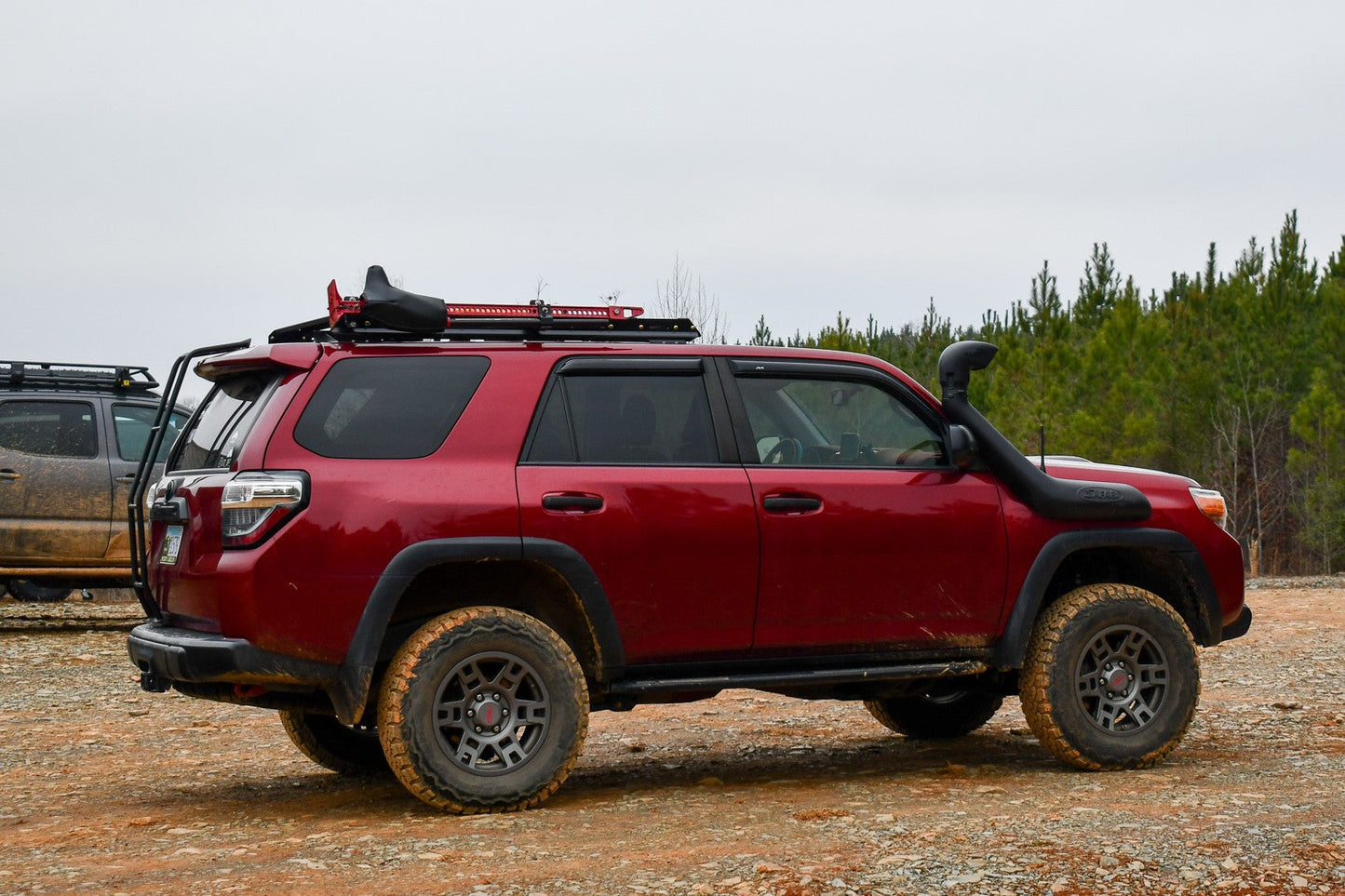 A 5th Gen 4Runner with a red finish, outfitted with off-road tires, a snorkel, and a 3/4 Roof Rack - 5th Gen 4Runner featuring ruggedized crossbars, sits slightly muddy on gravel near trees—hinting at recent off-road adventures.