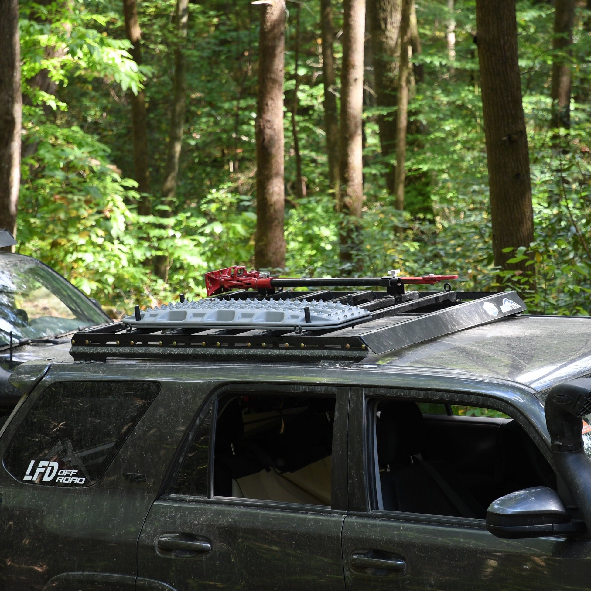 A dark SUV with LFD Off Road branding is parked in a forest, equipped with ruggedized crossbars on its 3/4 Roof Rack - 5th Gen 4Runner, loaded with recovery boards and roof rack accessories as sunlight filters through green trees.