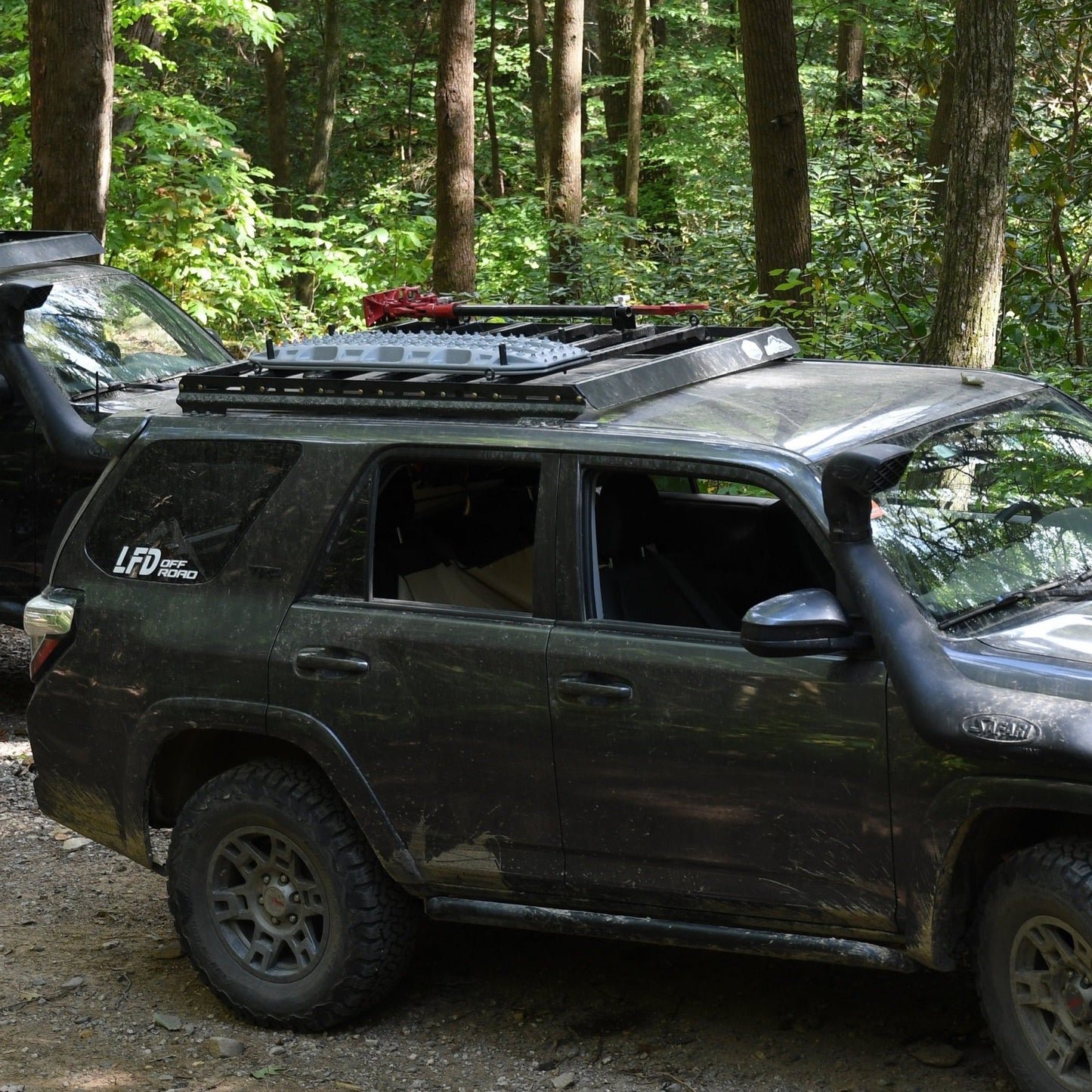 A 5th Gen 4Runner with a black 3/4 Roof Rack is parked on a dirt trail in a lush, green forest. Another similar SUV is partially visible behind it, surrounded by tall trees.