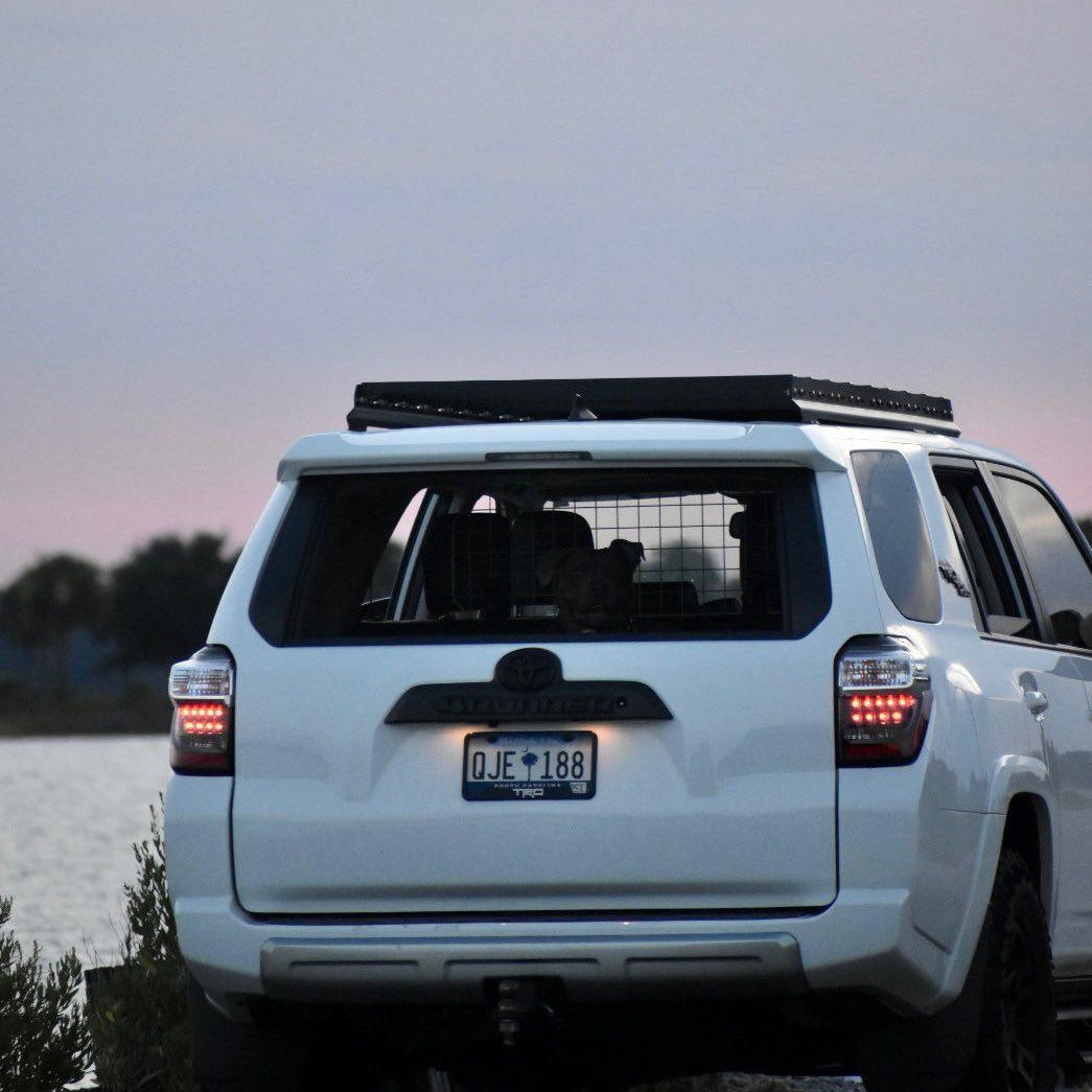 A white 5th Gen 4Runner with a 7/8 Roof Rack is parked by the water at dusk. The taillights are on, a dog in a metal crate is visible through the rear window, and trees frame a soft pink sky in the background.