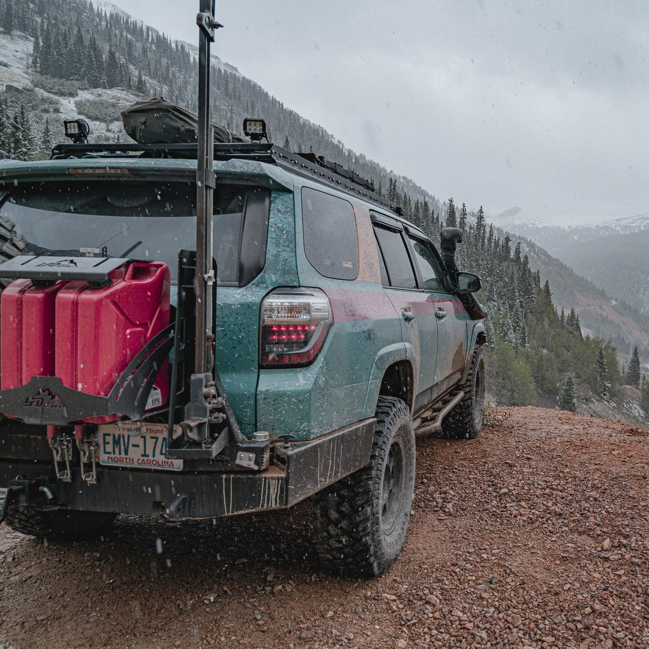 A rugged 5th Gen 4Runner with mud splatters, a rooftop tent, and extra gear mounted on the 7/8 Roof Rack - 5th Gen 4Runner is parked on a dirt road overlooking snow-covered, forested mountains.