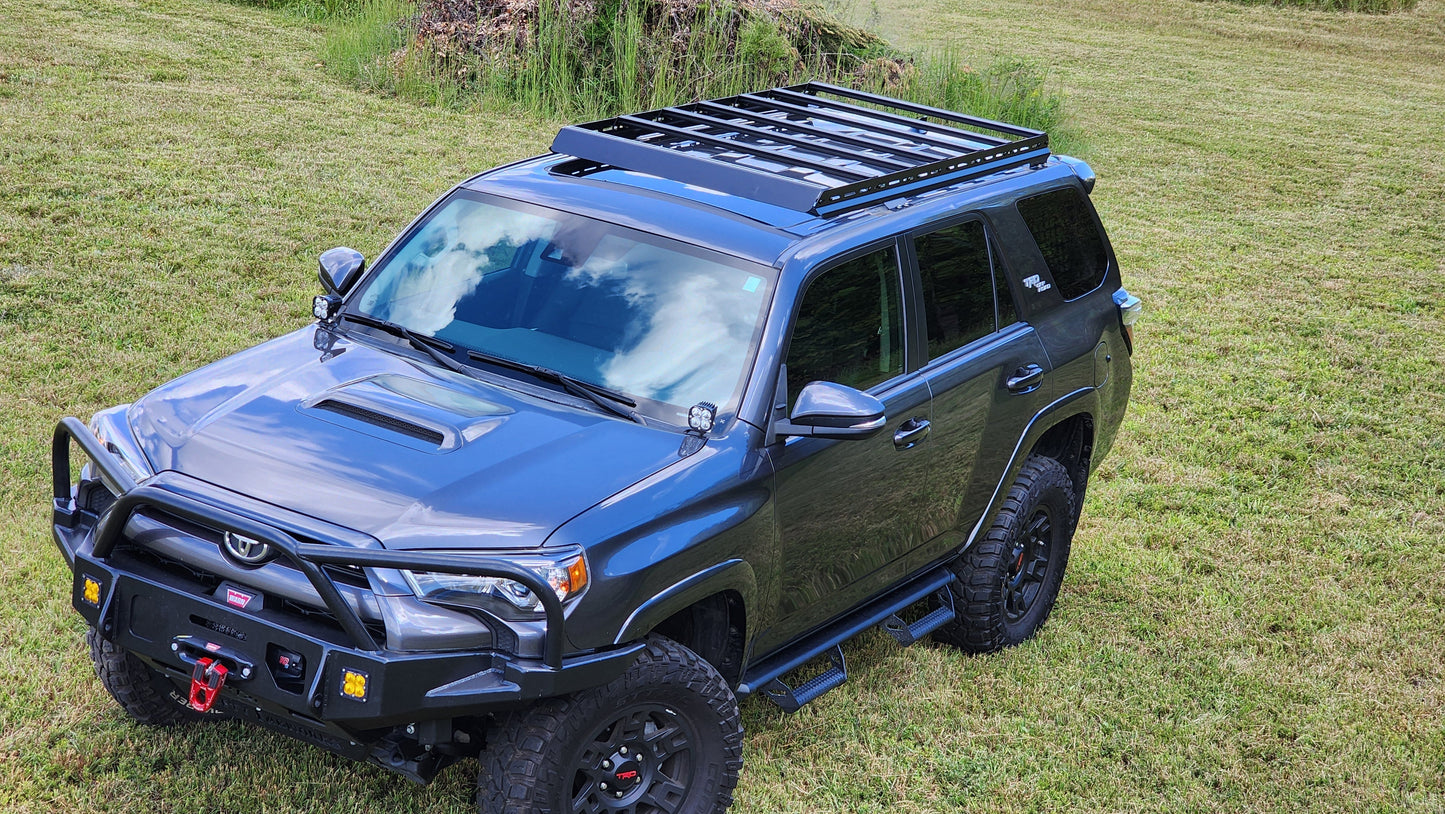 A 5th Gen 4Runner with a dark gray finish, equipped with a 7/8 Roof Rack featuring ruggedized crossbars, front bumper guard, and winch, is parked on green grass under a partly cloudy sky.