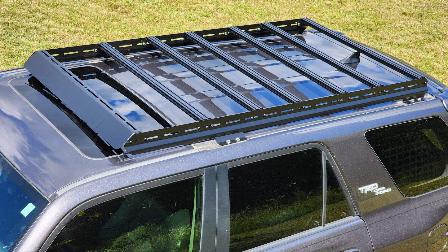 A 7/8 Roof Rack - 5th Gen 4Runner with ruggedized crossbars is mounted on a dark SUV parked on grass. The rack includes multiple slats and side rails, ideal for securing cargo or a rooftop tent.
