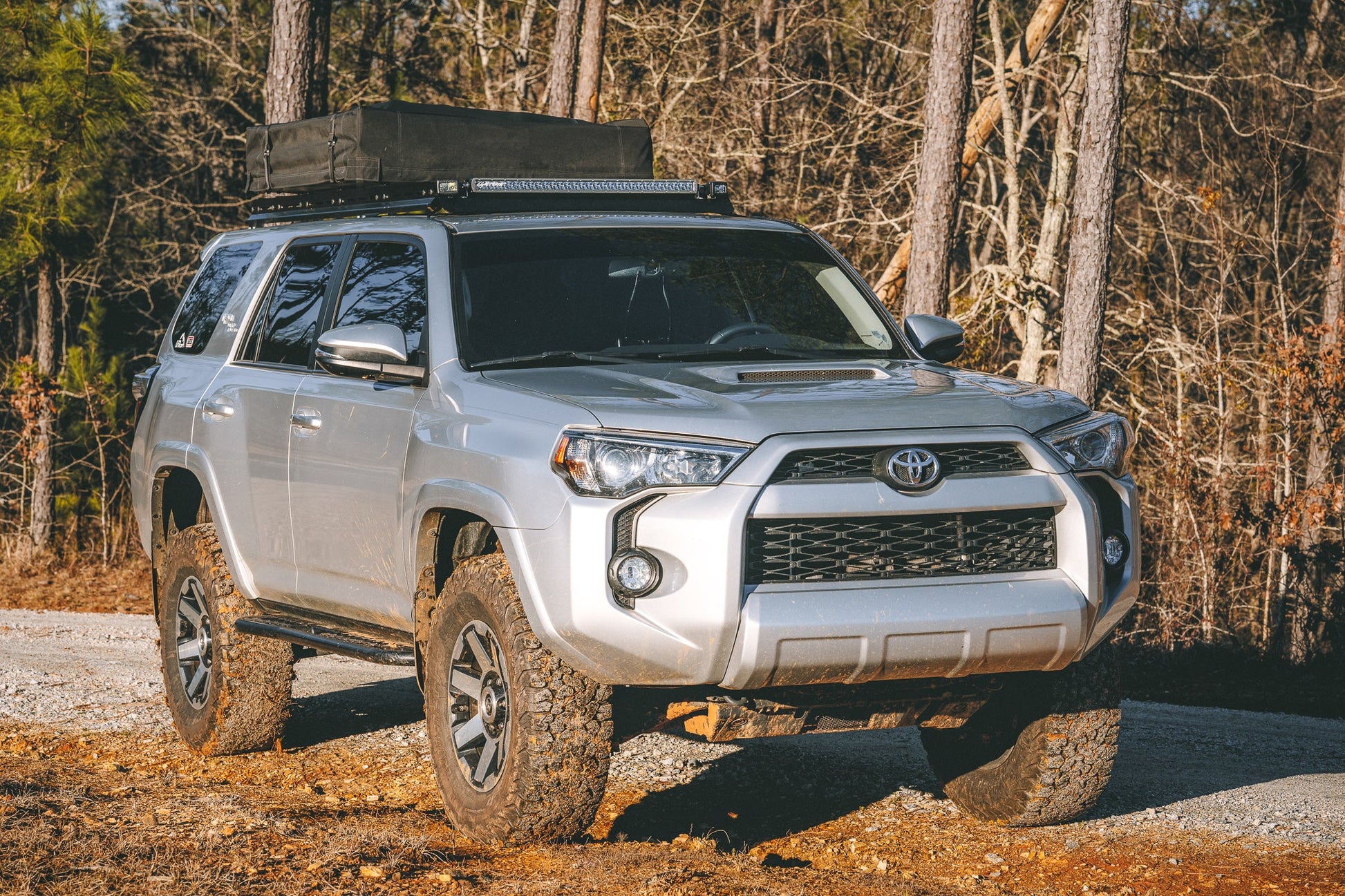 A silver Toyota 4Runner outfitted with the 7/8 Roof Rack - 5th Gen 4Runner and a roof cargo box is parked on a dirt road in the woods, mud splattered on its wheels and body.