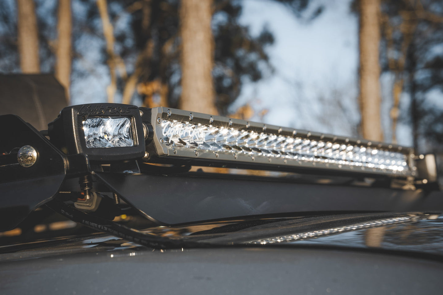 Close-up of a mounted LED light bar and spotlight on ruggedized crossbars atop the 7/8 Roof Rack - 5th Gen 4Runner, with trees and sky softly blurred in the background.