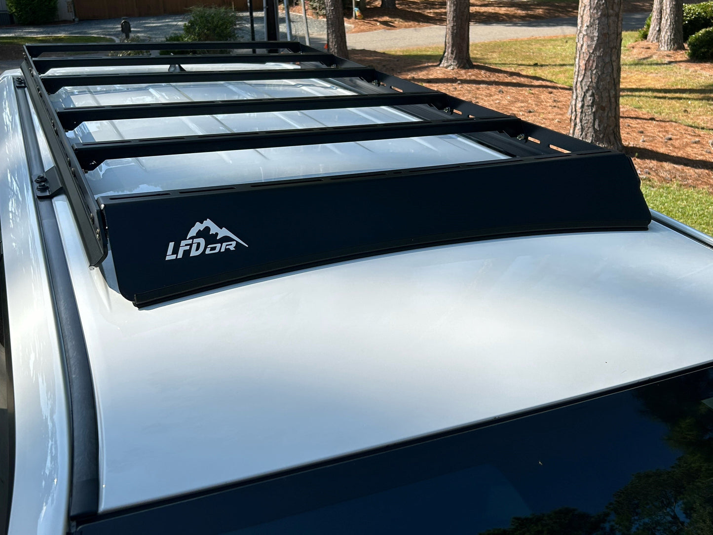 A close-up of a black 7/8 Roof Rack - 5th Gen 4Runner with rugged crossbars, mounted on a white vehicle and ideal for roof top tents. Trees and grass form the background.