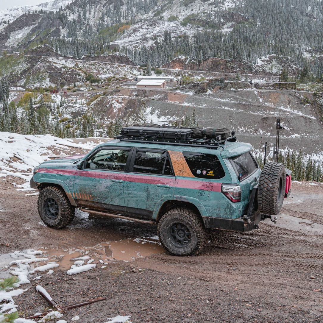 A teal 5th Gen 4Runner with off-road tires and a 7/8 Roof Rack is parked on a snowy mountain trail, roof gear secured by ruggedized crossbars. Snowy peaks and scattered trees are visible in the valley below.