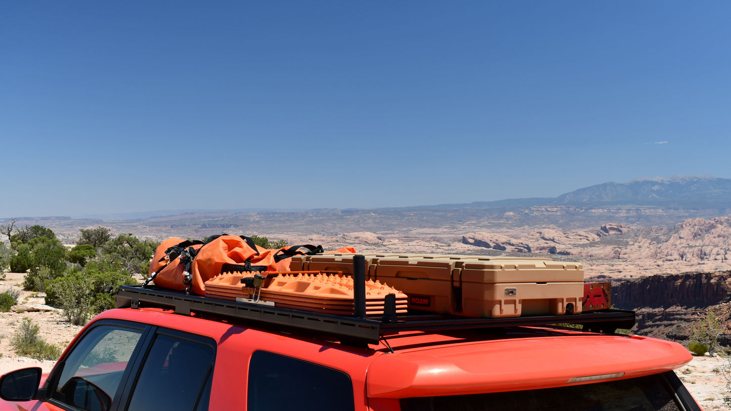 A 5th Gen 4Runner with a Ruggedized Low Profile Full Rack, loaded with outdoor gear and storage boxes, is parked overlooking a vast, rocky desert under a clear blue sky.