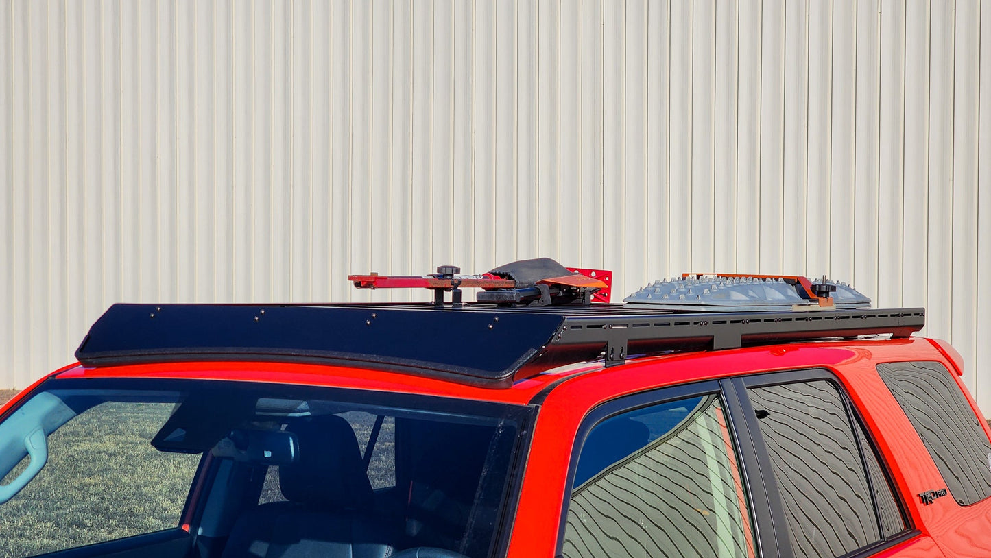 A red 5th Gen 4Runner with a black Ruggedized Low Profile Full Rack is parked by a beige corrugated metal wall. The rack carries traction boards and a shovel, showcasing its modular off-road roof rack system for tools and gear.