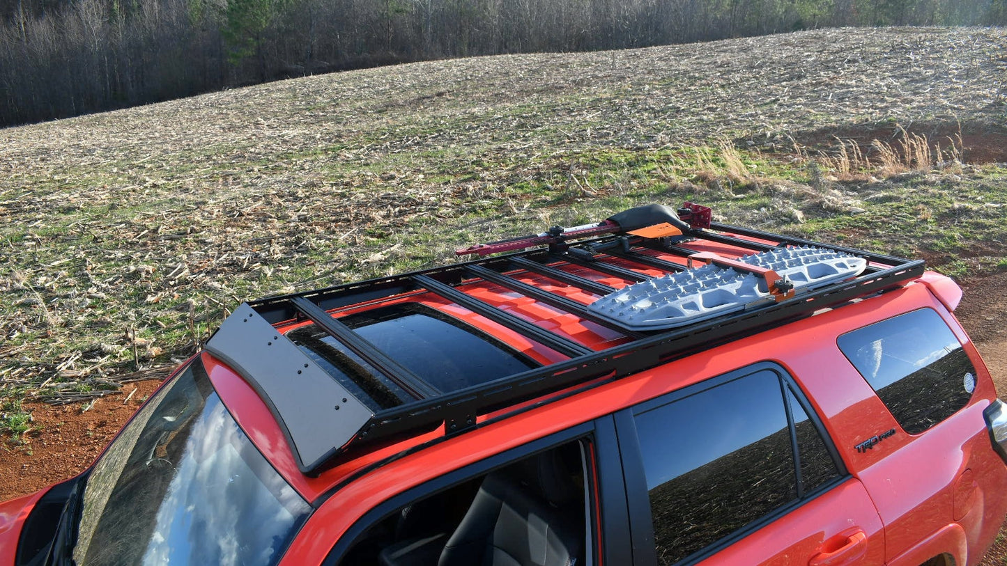 A red 5th Gen 4Runner with a Ruggedized Low Profile Full Rack is parked in a harvested rural field, equipped with heavy-duty crossbars holding a recovery board, ax, and off-road gear; trees are visible in the background.