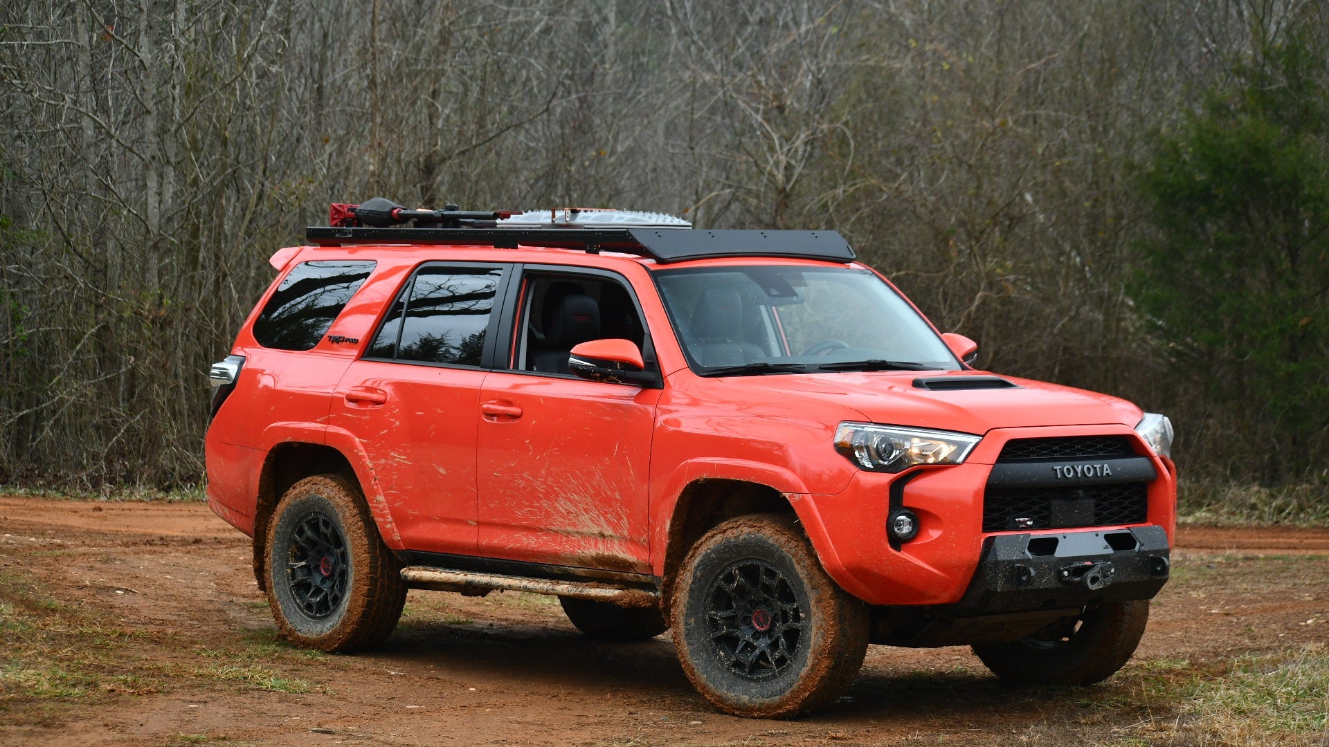 A red Toyota SUV with muddy tires and a Ruggedized Low Profile Full Rack - 5th Gen 4Runner is parked on a dirt trail in a forested area, surrounded by leafless trees and greenery.
