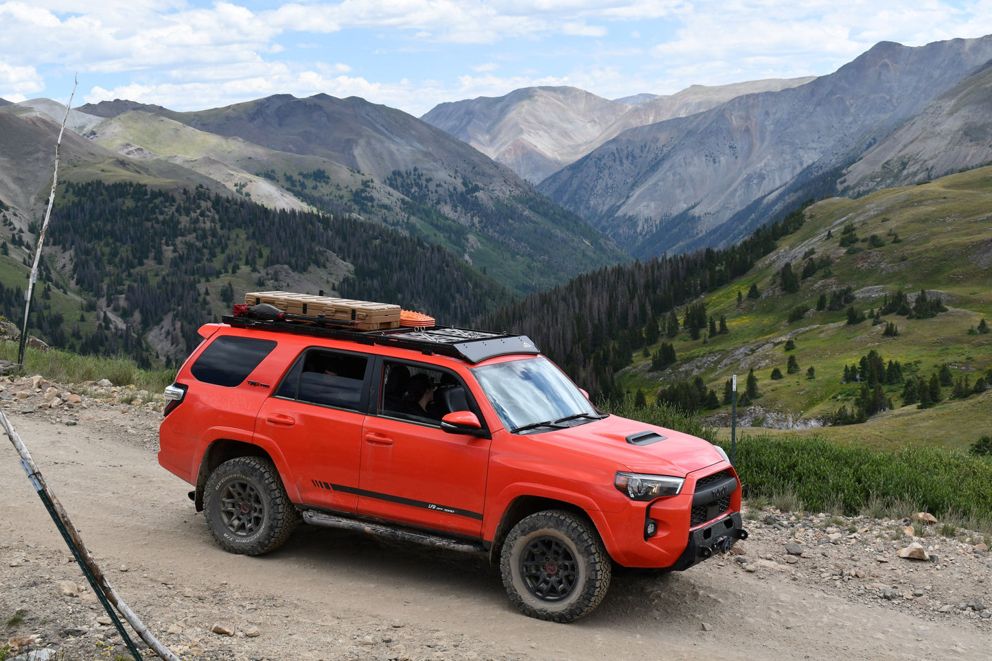 A 5th Gen 4Runner equipped with the Ruggedized Low Profile Full Rack, loaded with gear, tackles a rocky mountain trail amid green hills, trees, and distant peaks below a partly cloudy sky.