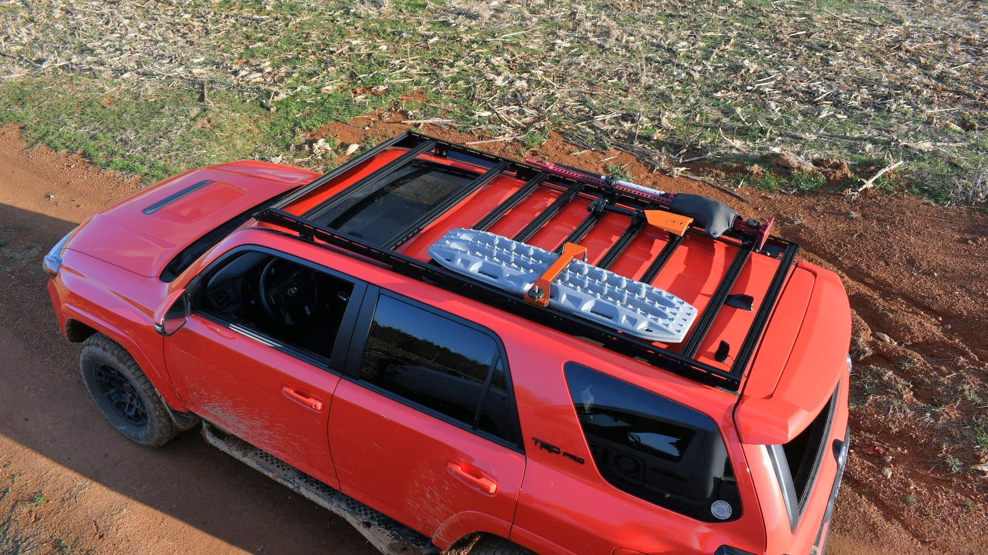 A 5th Gen 4Runner with a Ruggedized Low Profile Full Rack holds traction boards, a shovel, and gear as it’s parked on a dirt road beside a field of dry, cut vegetation.