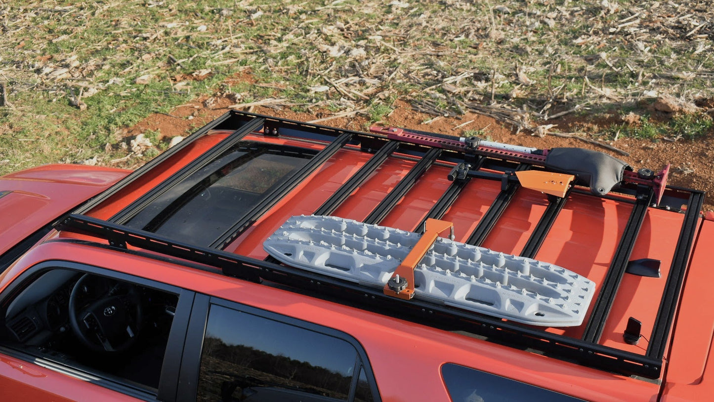 A red 5th Gen 4Runner with the Ruggedized Low Profile Full Rack is pictured from above, transporting a gray traction board and gear. The SUV is parked on grassy, uneven terrain with scattered tree debris.