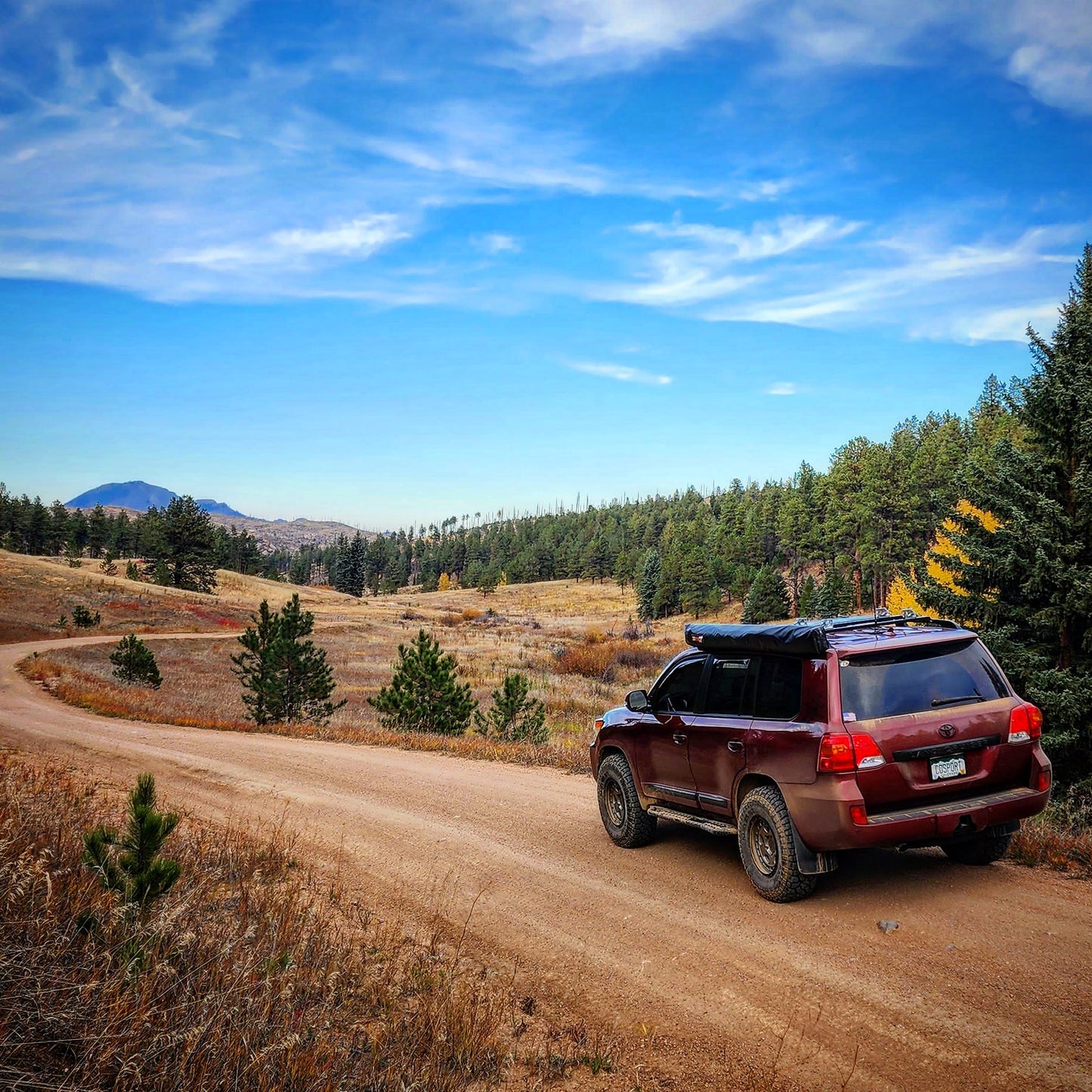A Land Cruiser 200 with a Ruggedized Crossbar Kit drives on a winding dirt road, surrounded by pine trees, grassy fields, and distant mountains beneath a blue sky with wispy clouds.