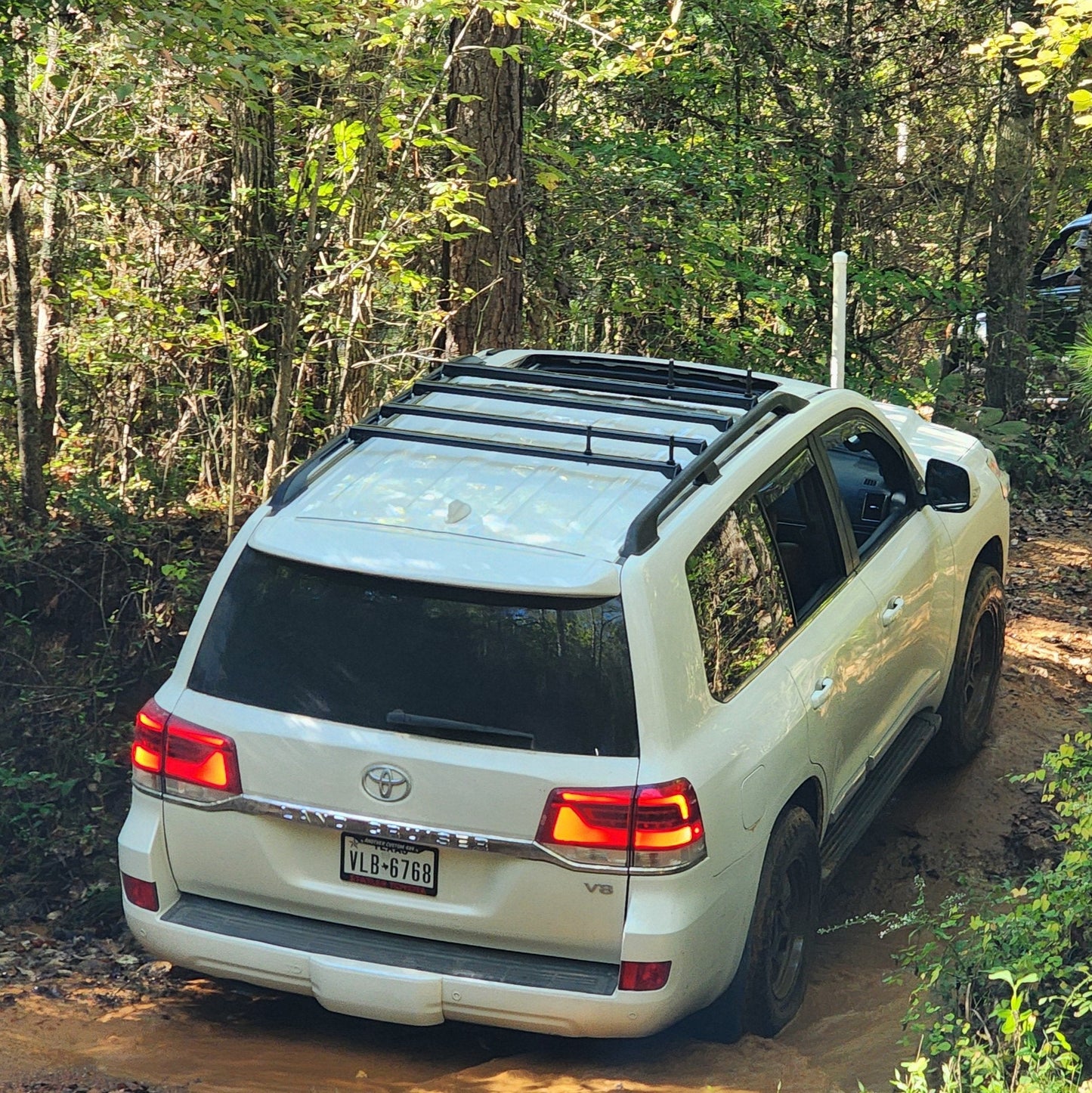 A white Land Cruiser 200 with the Ruggedized Crossbar Kit - Land Cruiser 200 navigates a muddy, narrow trail in a dense green forest as sunlight filters through the trees, highlighting its adventurous route.