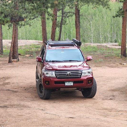 A maroon Land Cruiser 200 with off-road tires is parked on sand among pines. Outfitted with the Ruggedized Crossbar Kit - Land Cruiser 200 and a Colorado plate, this SUV stands ready for any adventure.