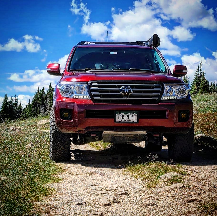 A Land Cruiser 200 equipped with the Ruggedized Crossbar Kit is parked on a rocky dirt trail, surrounded by grass and trees under a blue sky. The license plate reads COSPORT.