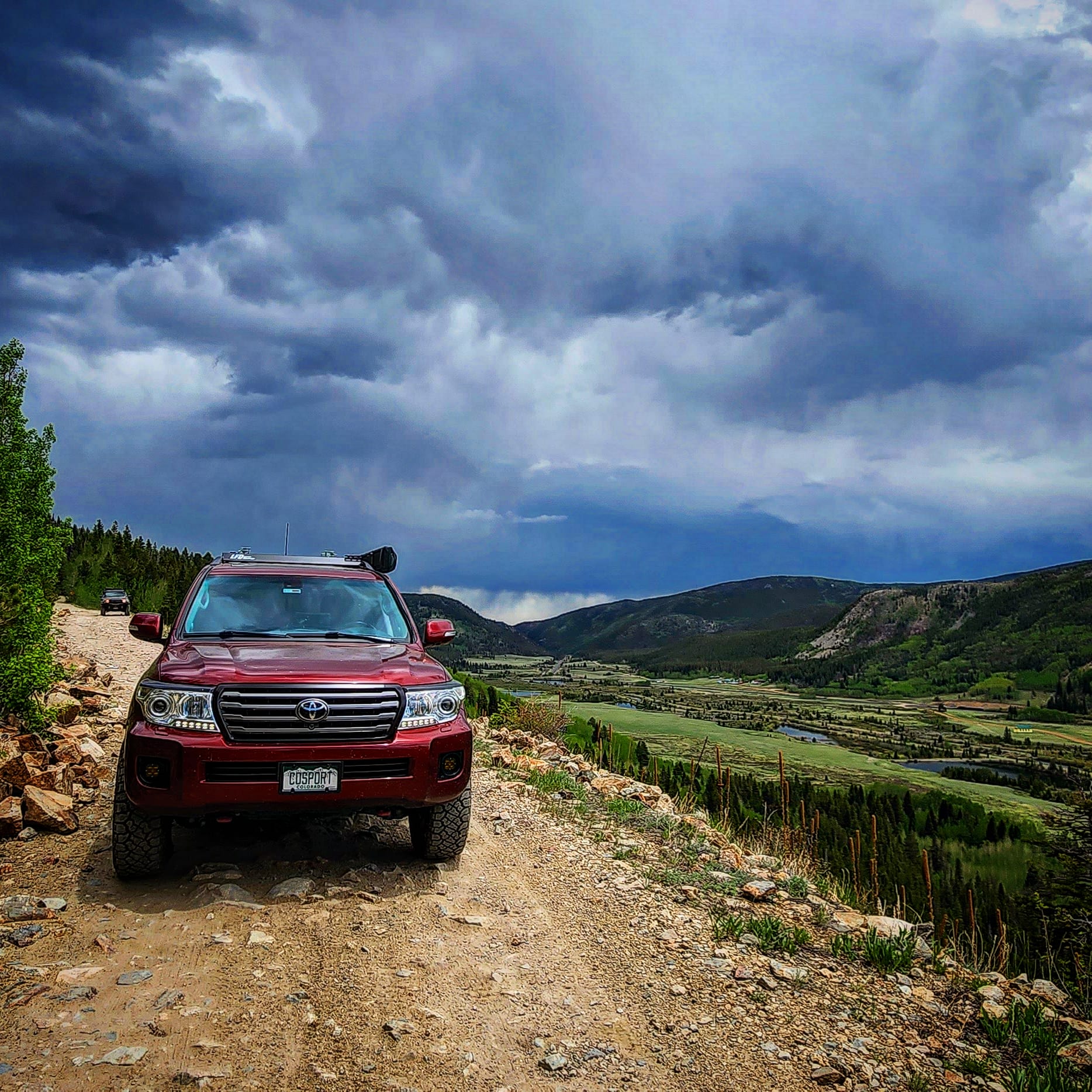 A Land Cruiser 200 equipped with the Ruggedized Crossbar Kit is parked on a rocky dirt road overlooking a green valley and distant mountains under dramatic clouds. Another vehicle can be seen further down the road.