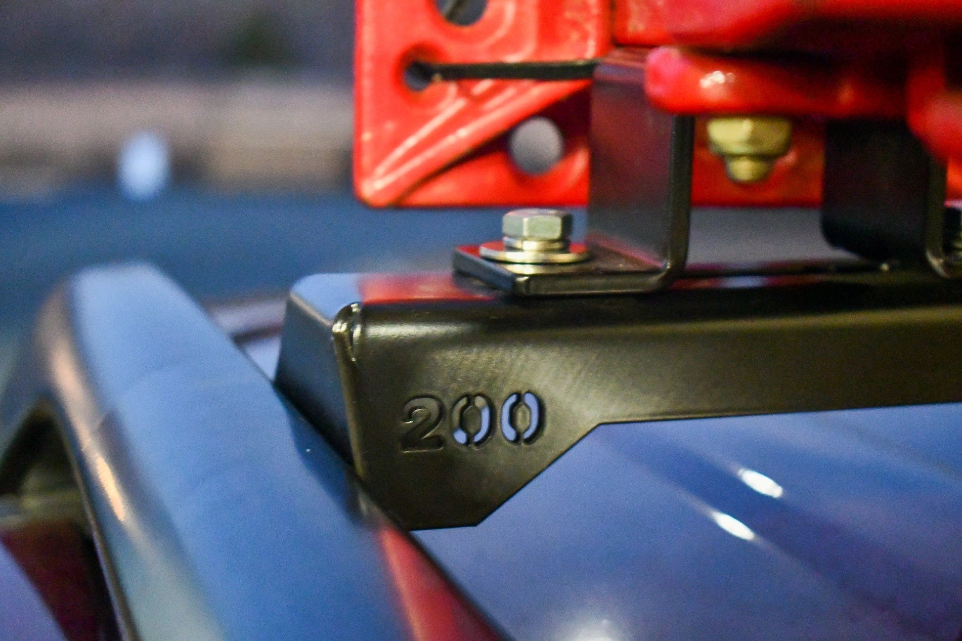 Close-up of a metal bracket engraved with “200,” attached by bolts to a red part on Land Cruiser 200 roof rails—part of the Ruggedized Crossbar Kit - Land Cruiser 200 for enhanced durability.