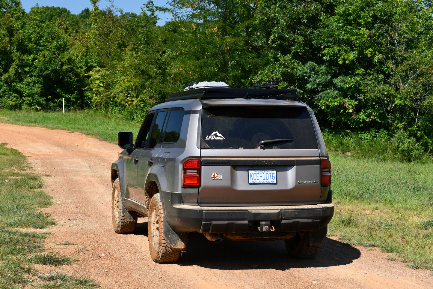 A gray SUV with muddy tires drives on a dirt road flanked by grass and trees under a sunny sky. It’s equipped with the 7/8 Roof Rack - 250 Series Land Cruiser (2024+), featuring rugged crossbars and a visible rear license plate.