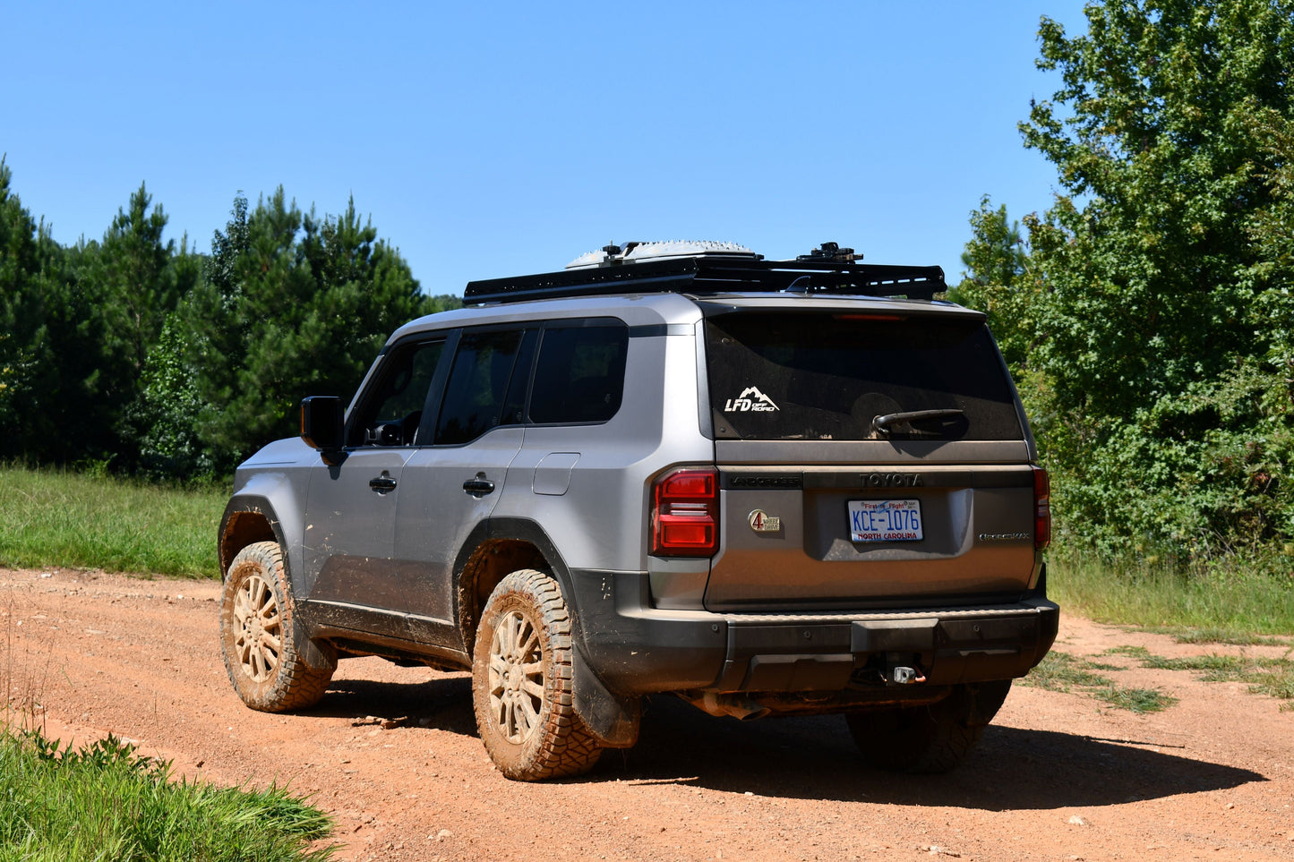 A gray 2024+ Toyota Land Cruiser 250 Series with muddy tires is parked on a dirt road, featuring rugged crossbars atop a 7/8 Roof Rack, surrounded by green grass and trees beneath a clear blue sky.