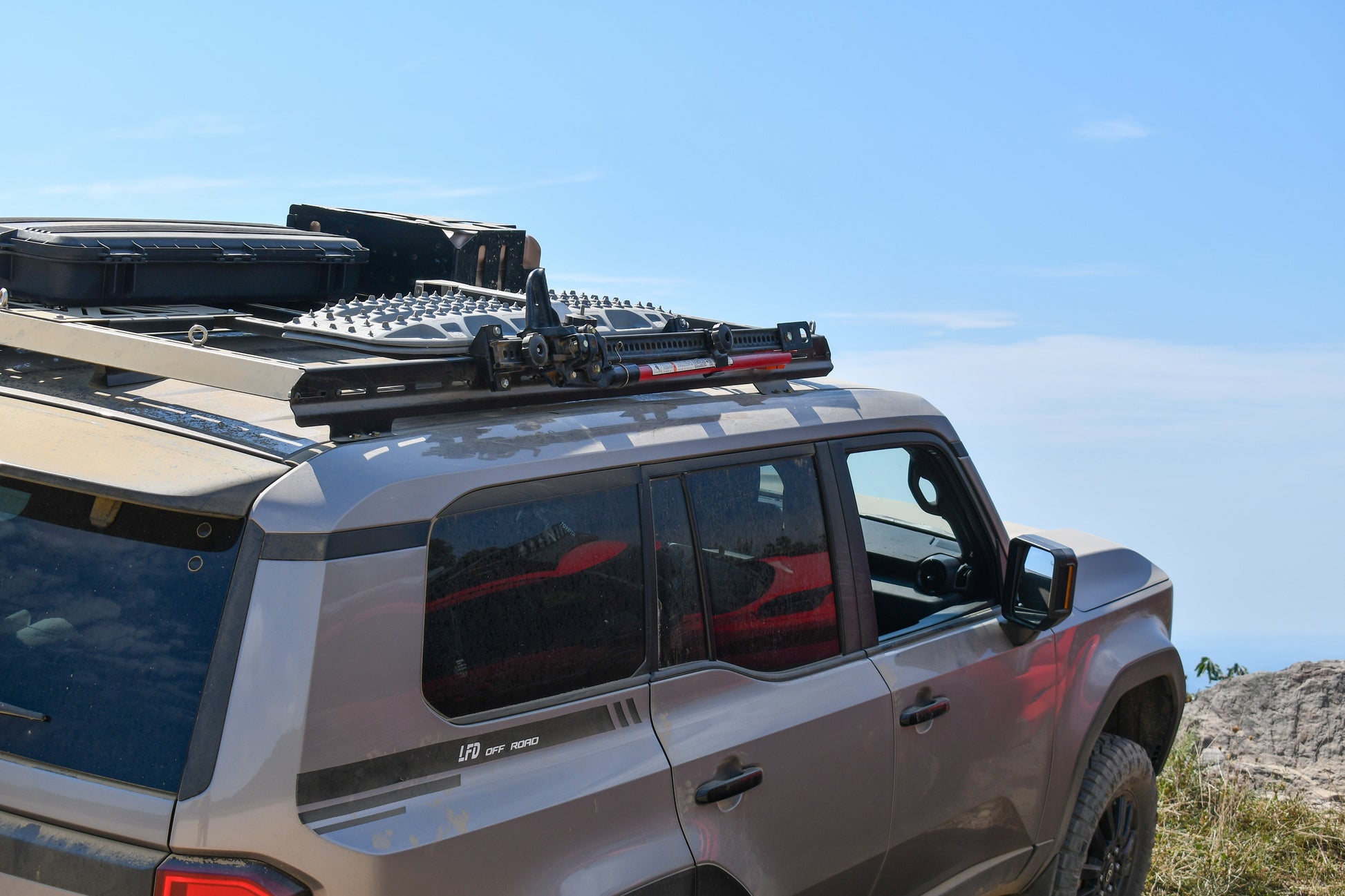 A tan off-road SUV sits under a blue sky, featuring the 7/8 Roof Rack - 250 Series Land Cruiser (2024+) with rugged crossbars carrying a storage box, traction boards, and a jack—ready for any adventure.