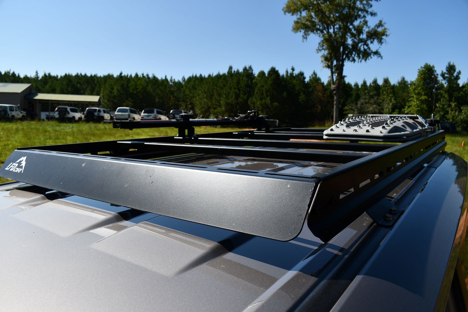 A close-up of the 7/8 Roof Rack - 250 Series Land Cruiser (2024+) rugged crossbars mounted on a silver vehicle, with trees, parked cars, and a building visible in the sunny background.
