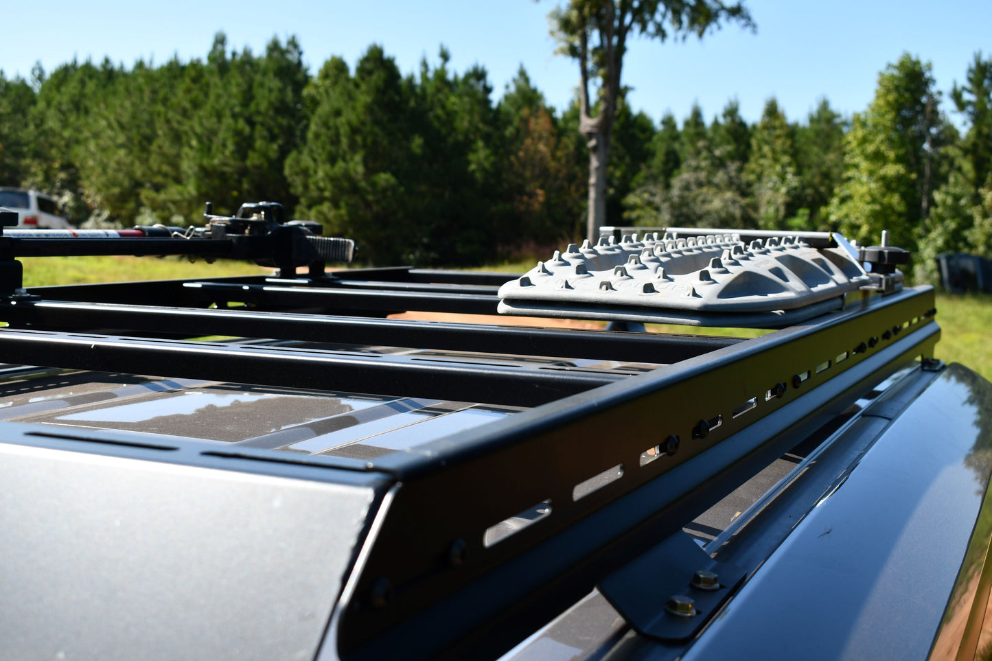 Close-up of the 7/8 Roof Rack - 250 Series Land Cruiser (2024+) featuring ruggedized crossbars and recovery traction boards, mounted on a Land Cruiser parked outdoors by grass and a forested backdrop on a sunny day.