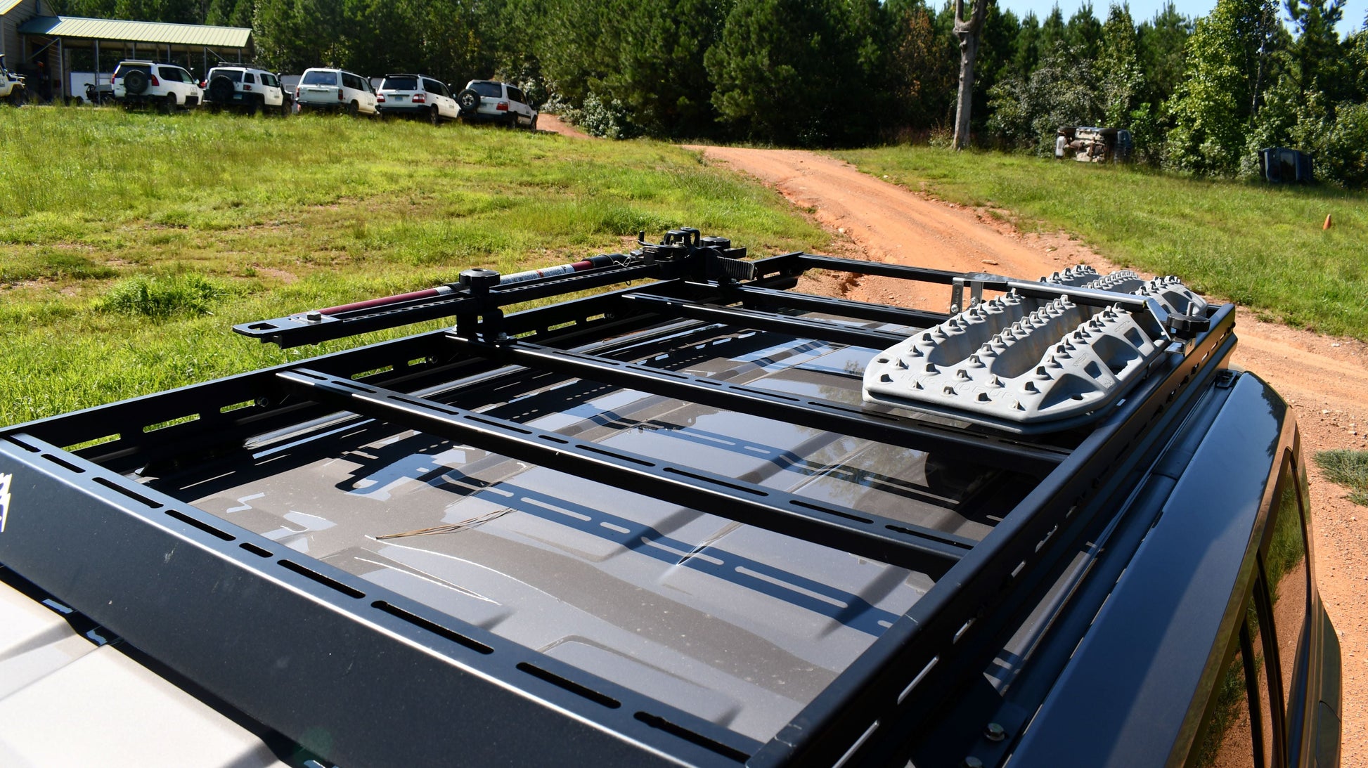 A 7/8 Roof Rack - 250 Series Land Cruiser (2024+) with ruggedized crossbars and recovery boards is mounted on a vehicle parked on a dirt road, surrounded by grass, trees, and several white SUVs in the background.