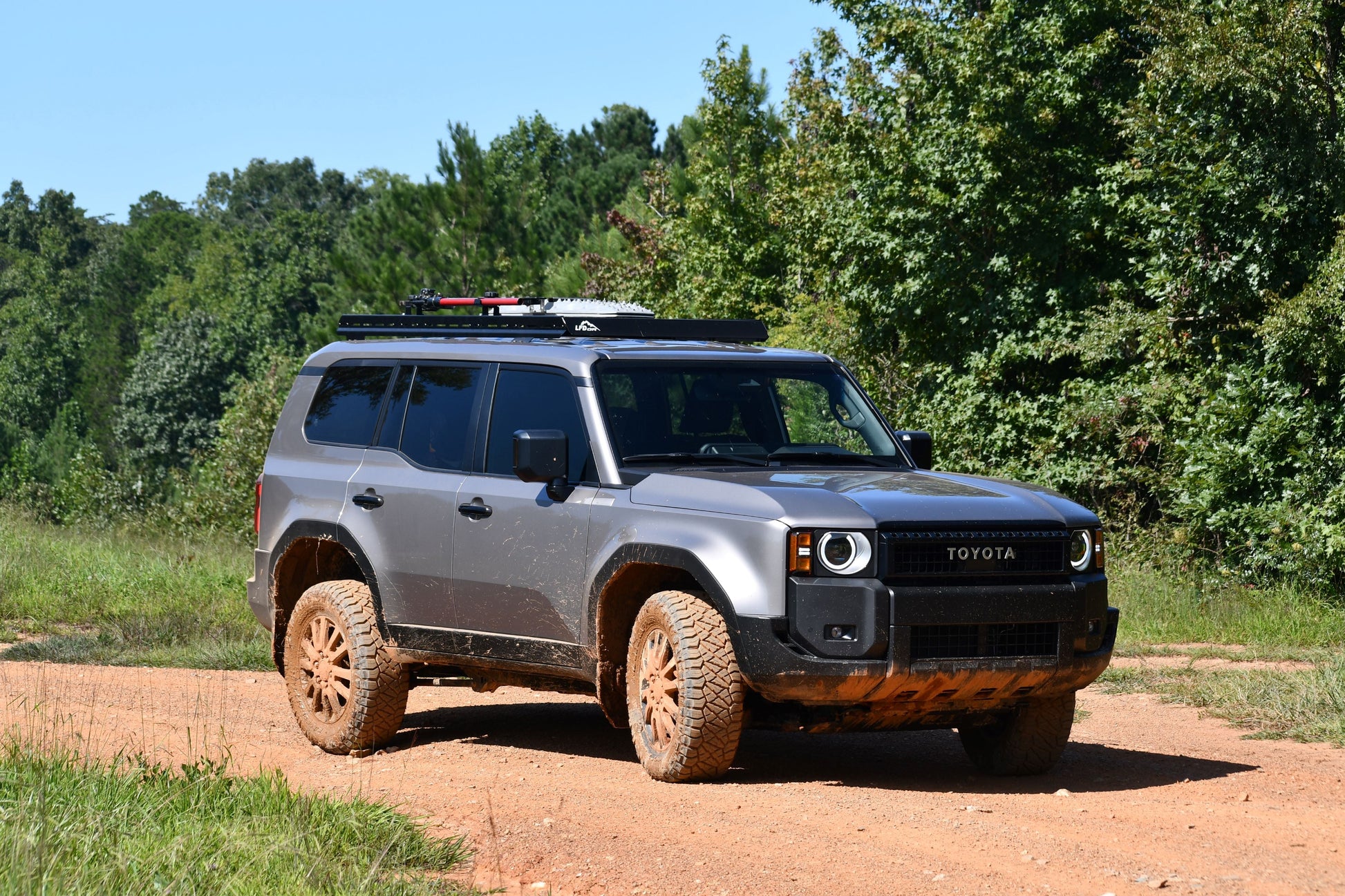 A gray 2024+ Toyota Land Cruiser 250 Series with muddy tires and a 7/8 Roof Rack is parked on a dirt road surrounded by green trees and grass under a clear sky.