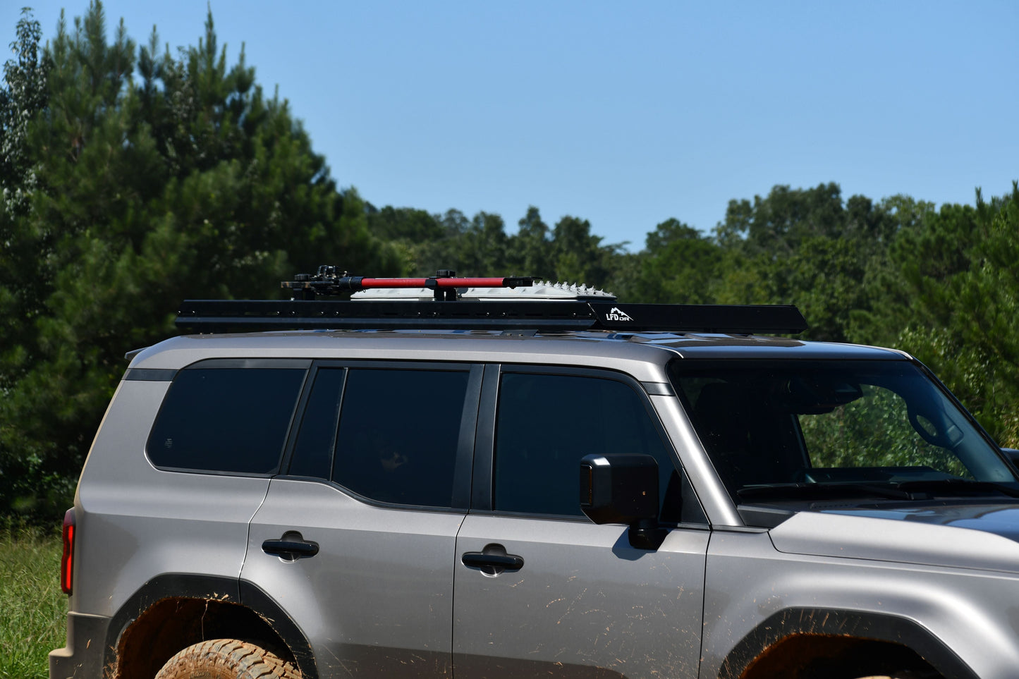 A 2024+ 250 Series Land Cruiser with a 7/8 Roof Rack is parked outdoors on a sunny day, carrying off-road gear and surrounded by green trees and grass.