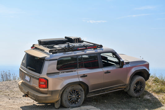 A tan 2024+ 250 Series Land Cruiser with off-road tires and a 7/8 Roof Rack is loaded with camping gear and parked on a dirt trail, overlooking a scenic, hazy landscape under a clear sky.