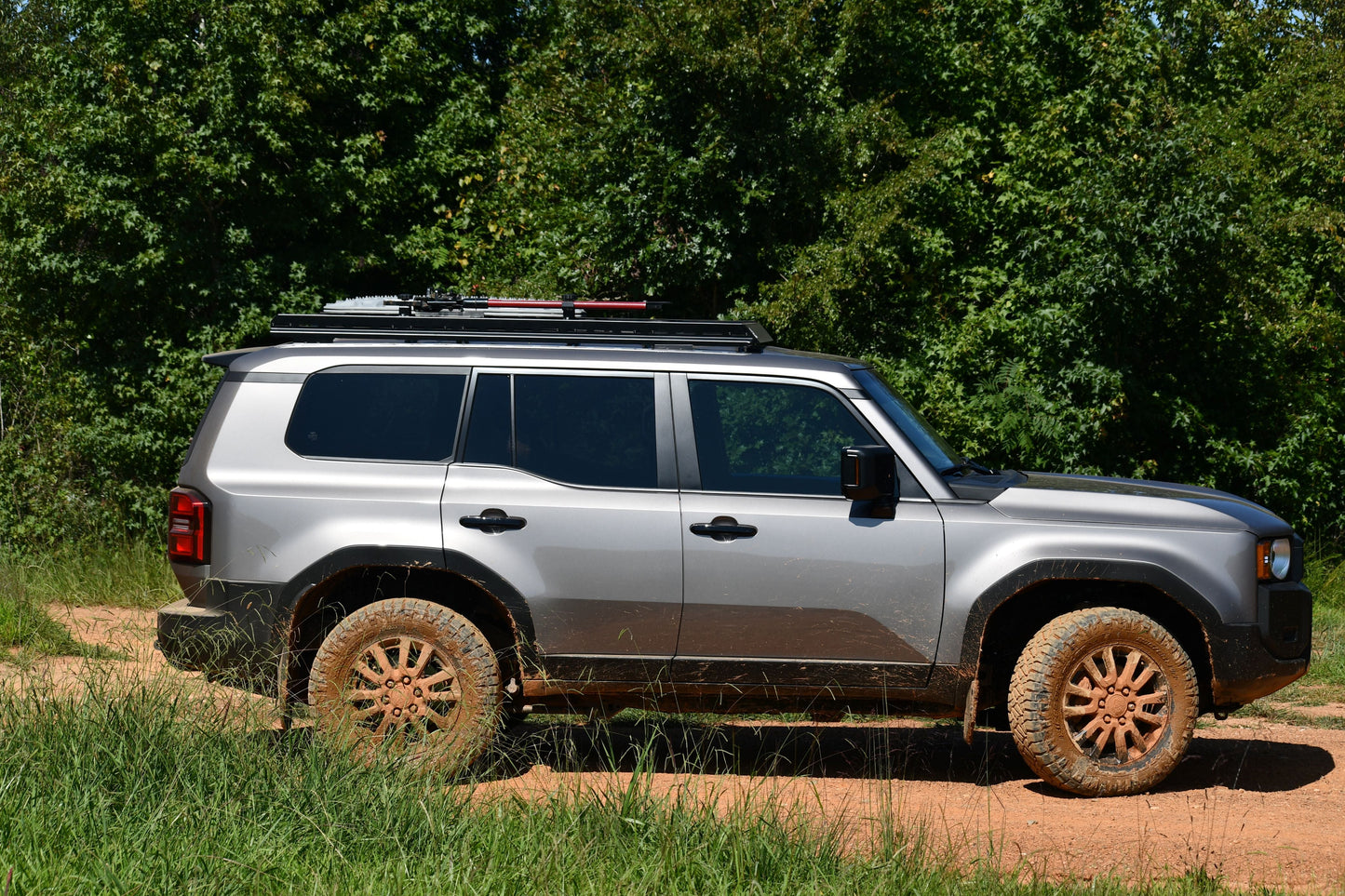 A silver 2024+ 250 Series Land Cruiser with muddy tires and a 7/8 Roof Rack is parked on a dirt road amid green trees and grass, shown in side profile with ruggedized crossbars for adventure-ready style.