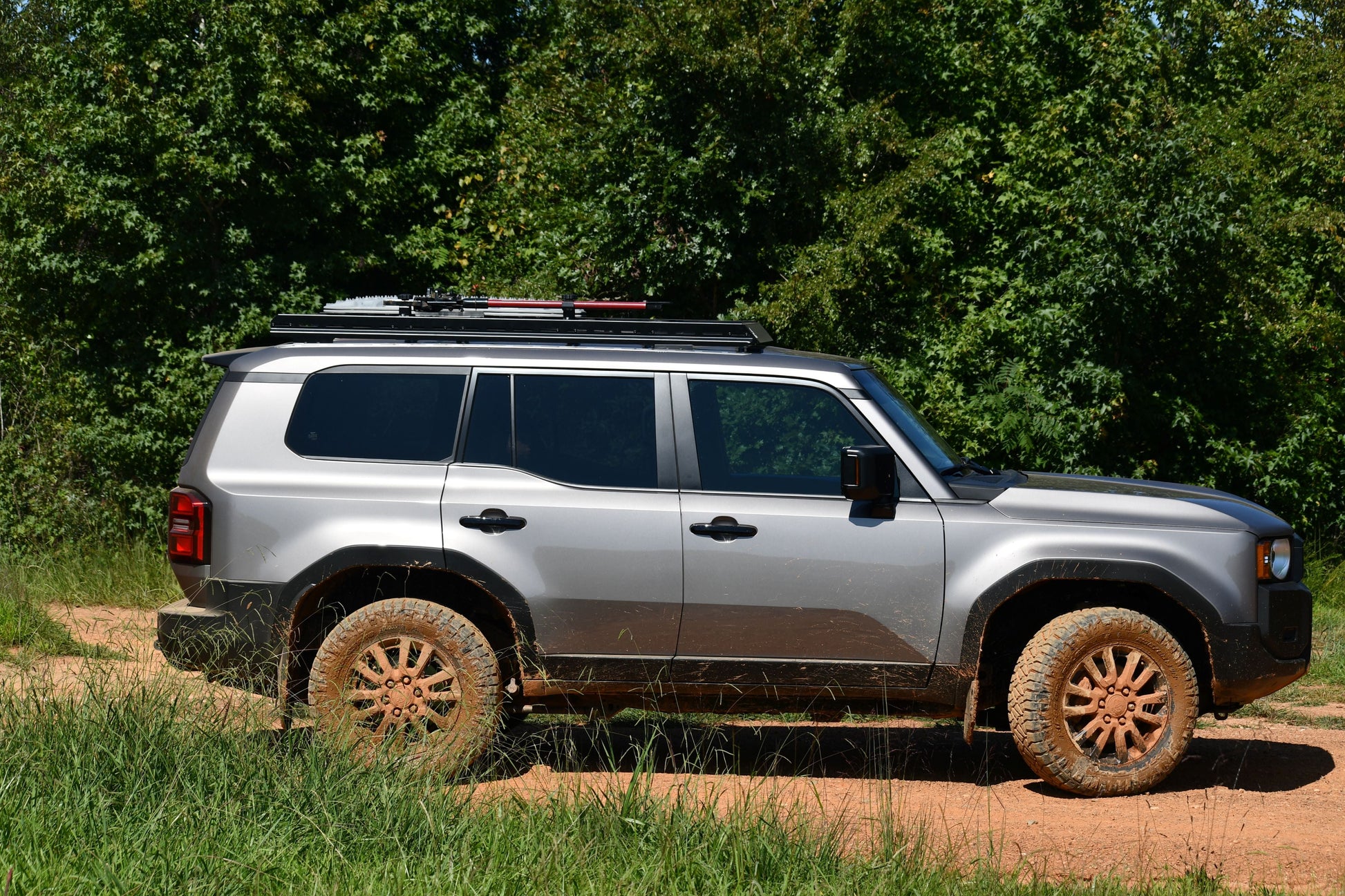 A silver 2024+ 250 Series Land Cruiser with muddy tires and a 7/8 Roof Rack is parked on a dirt road amid green trees and grass, shown in side profile with ruggedized crossbars for adventure-ready style.