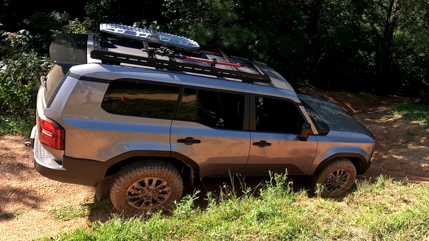 A 2024+ 250 Series Land Cruiser with a 7/8 Roof Rack drives up a dirt trail, muddy tires spinning as it climbs through lush greenery and trees.