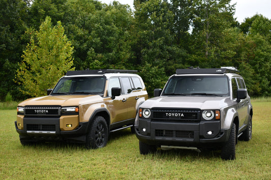 Two Toyota SUVs, one tan and one gray, are parked side by side on grass with trees behind them. Both feature rugged looks and are equipped with Ruggedized Aluminum Low Profile Rack - 250 Series Land Cruiser (2024+) for extra versatility.