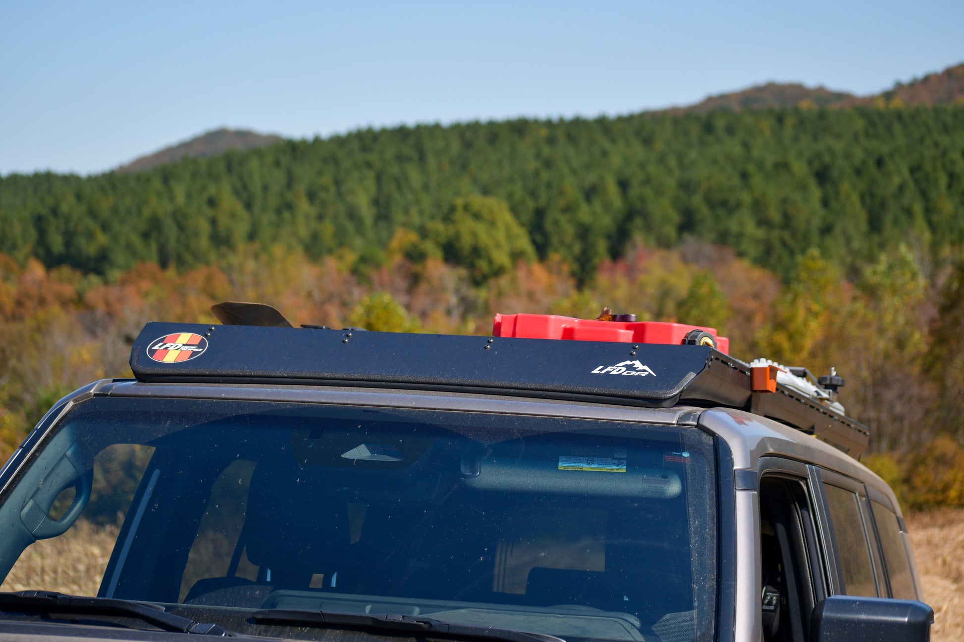 A Ruggedized Aluminum Low Profile Rack for the 2024+ 250 Series Land Cruiser, seen with modular crossbars holding gear and a red fuel canister, is parked before a scenic forested hillside under clear skies.