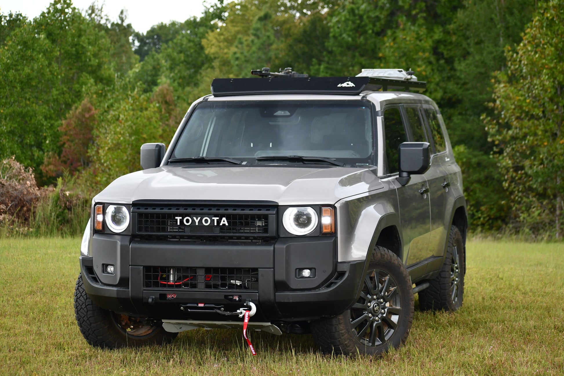 A gray 2024+ Toyota Land Cruiser 250 Series with a Ruggedized Aluminum Low Profile Rack is parked on grass before trees, featuring off-road tires, a winch, and a red tow hook on the front bumper.