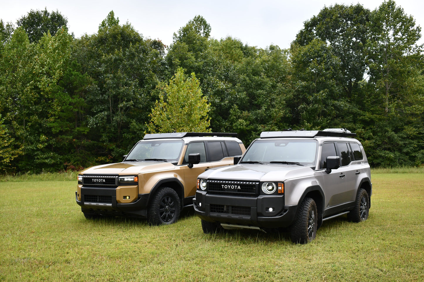 Two 2024+ Toyota Land Cruiser SUVs, one tan and one gray, are parked on grass with trees behind. Both feature rugged designs and are equipped with Ruggedized Aluminum Low Profile Racks for the 250 Series on their roof racks, facing slightly left.